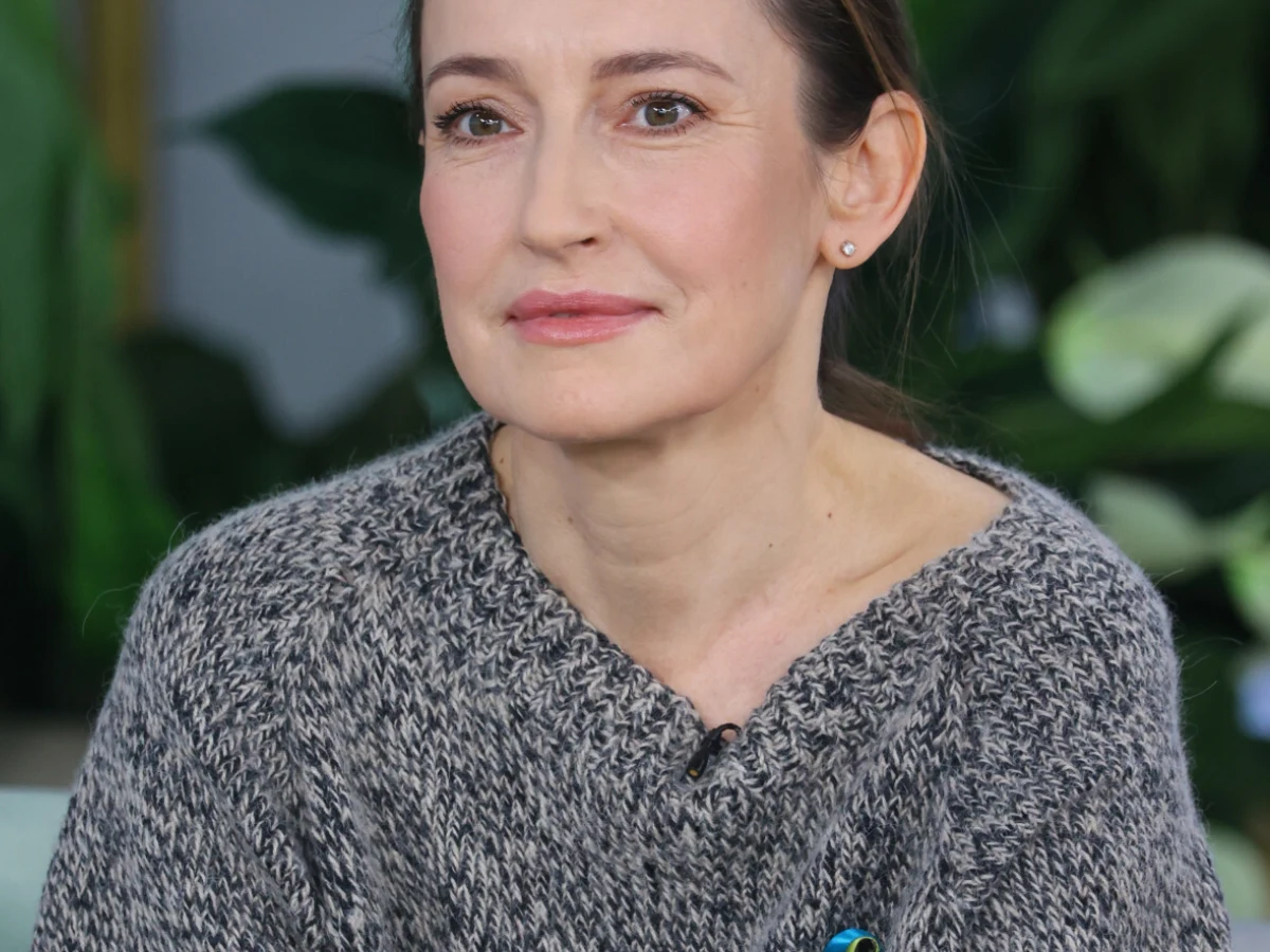 Katarzyna Groniec Katarzyna Groniec w szarym swetrze, pozująca na tle zielonych roślin.