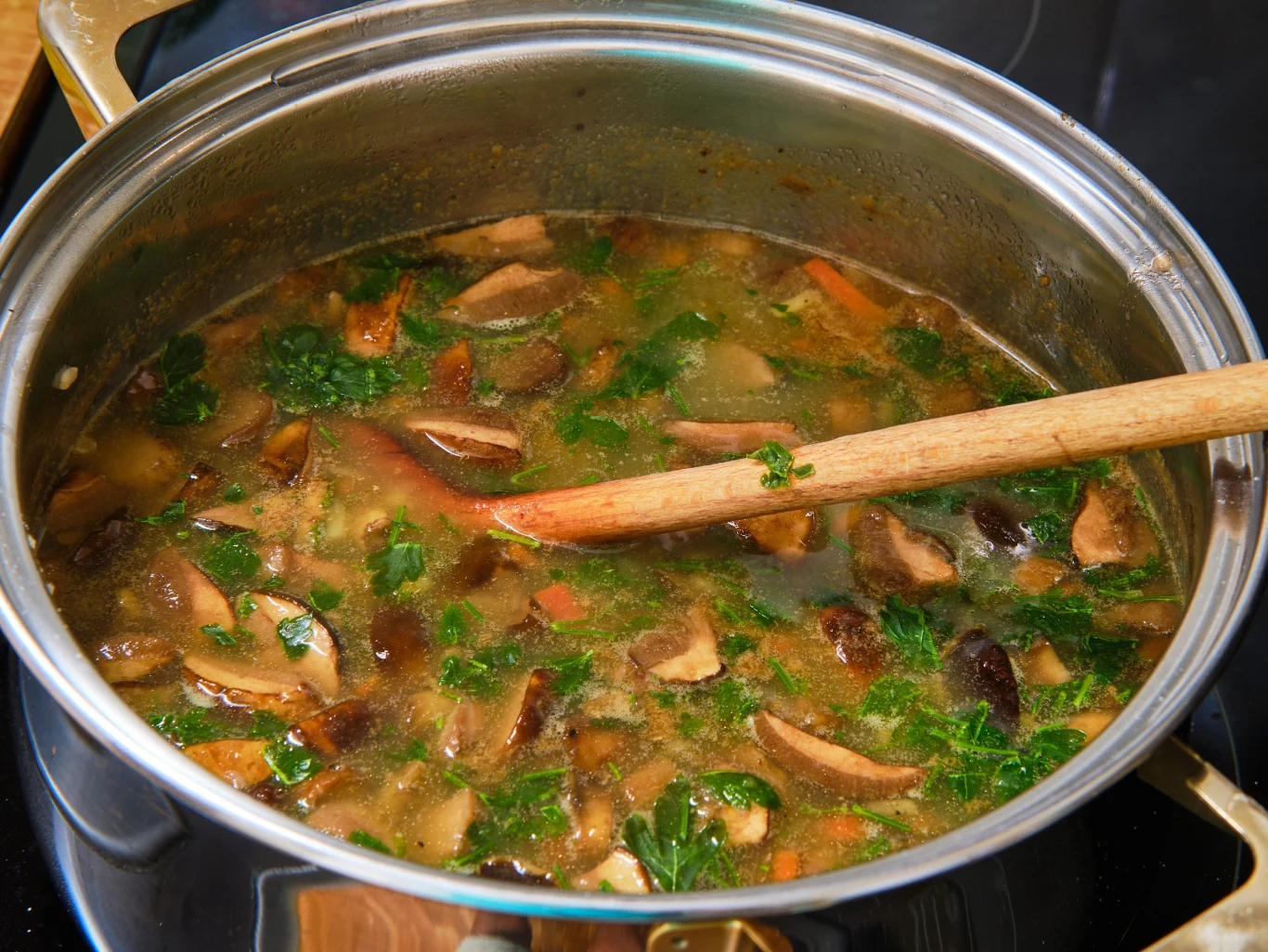 Zupa pieczarkowa z grzybami leśnymi to wymarzony jesienny obiad. Garnek z zupą pieczarkowo-grzybową, widoczne pokrojone grzyby, natka pietruszki i marchewka, mieszany drewnianą łyżką.