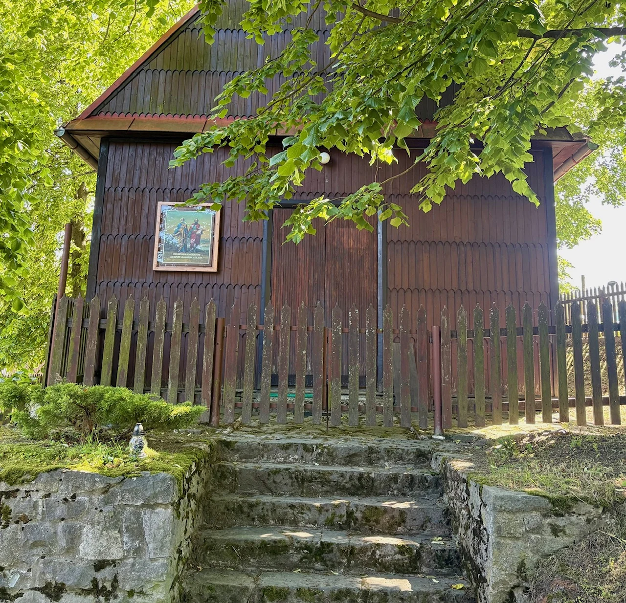 Drewniany budynek z wysokim ogrodzeniem i schodami z kamienia prowadzącymi wprost do zamkniętych drzwi, nad którymi wisi religijny obraz, otoczony zielenią i cieniem dużych drzew.