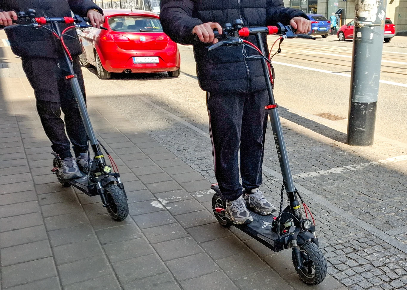 Zasady bezpiecznej jazdy hulajnogą. Policjanci z Legionowa przypominają obowiązujące przepisy. Dwie osoby jadące obok siebie na elektrycznych hulajnogach po chodniku w mieście, kontekst bezpiecznej jazdy.