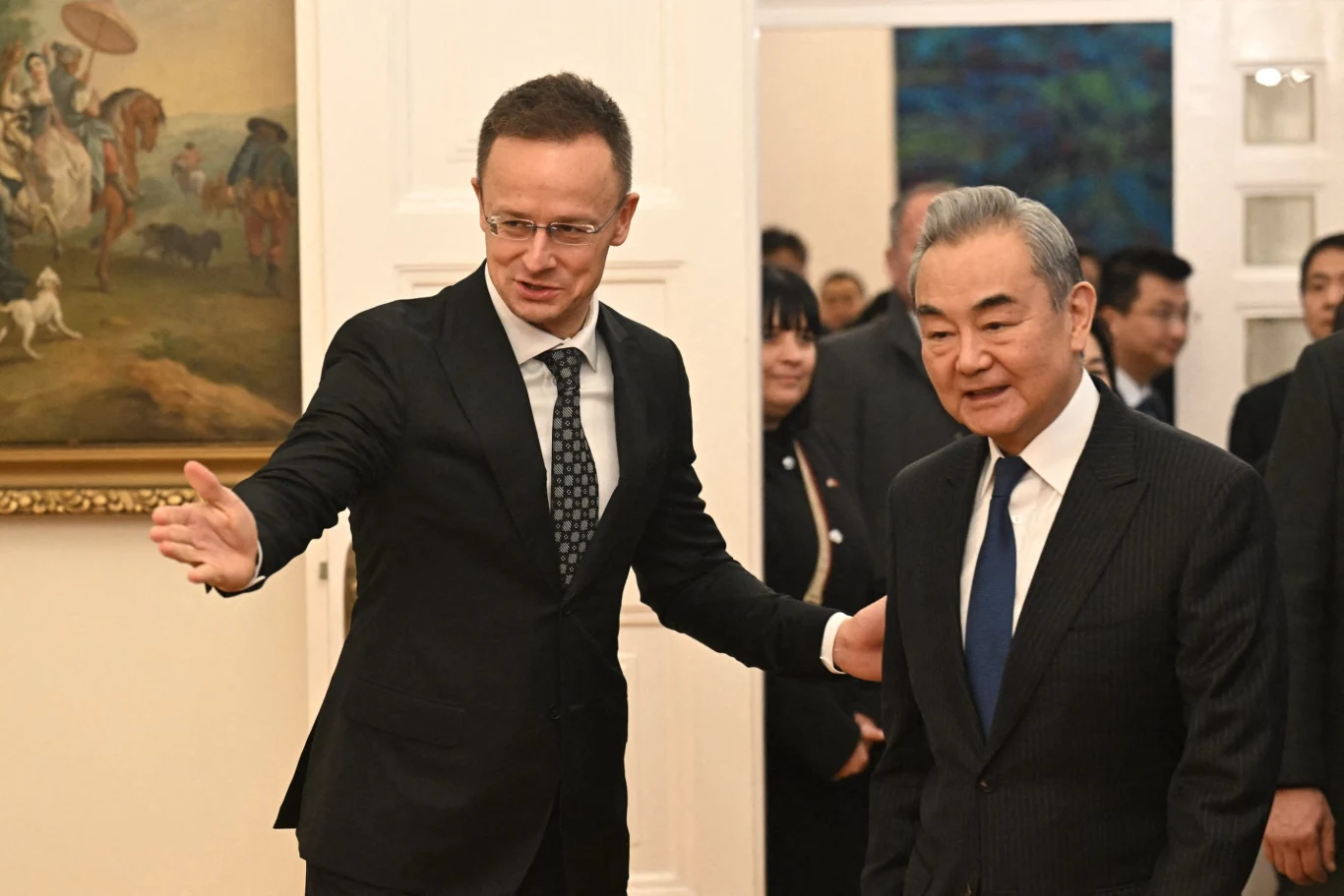 Dwóch elegancko ubranych mężczyzn w garniturach wymienia gesty podczas oficjalnego spotkania w eleganckim wnętrzu z obrazami na ścianach.