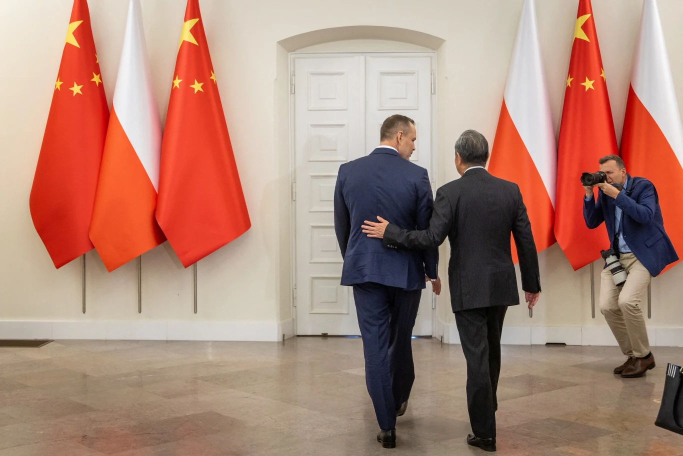 Dwóch mężczyzn w garniturach idzie korytarzem obok siebie, otoczonych flagami Polski i Chin, podczas gdy fotograf uwiecznia tę scenę.