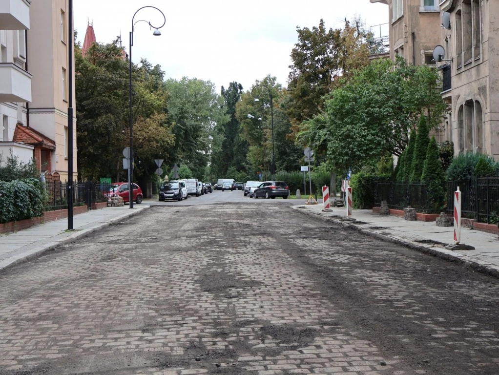 Remont ul. Roosevelta. Jakie utrudnienia czekają kierowców? Remont ul. Roosevelta w Legnicy: nierówna nawierzchnia, barierki ostrzegawcze, zaparkowane auta i budynki.