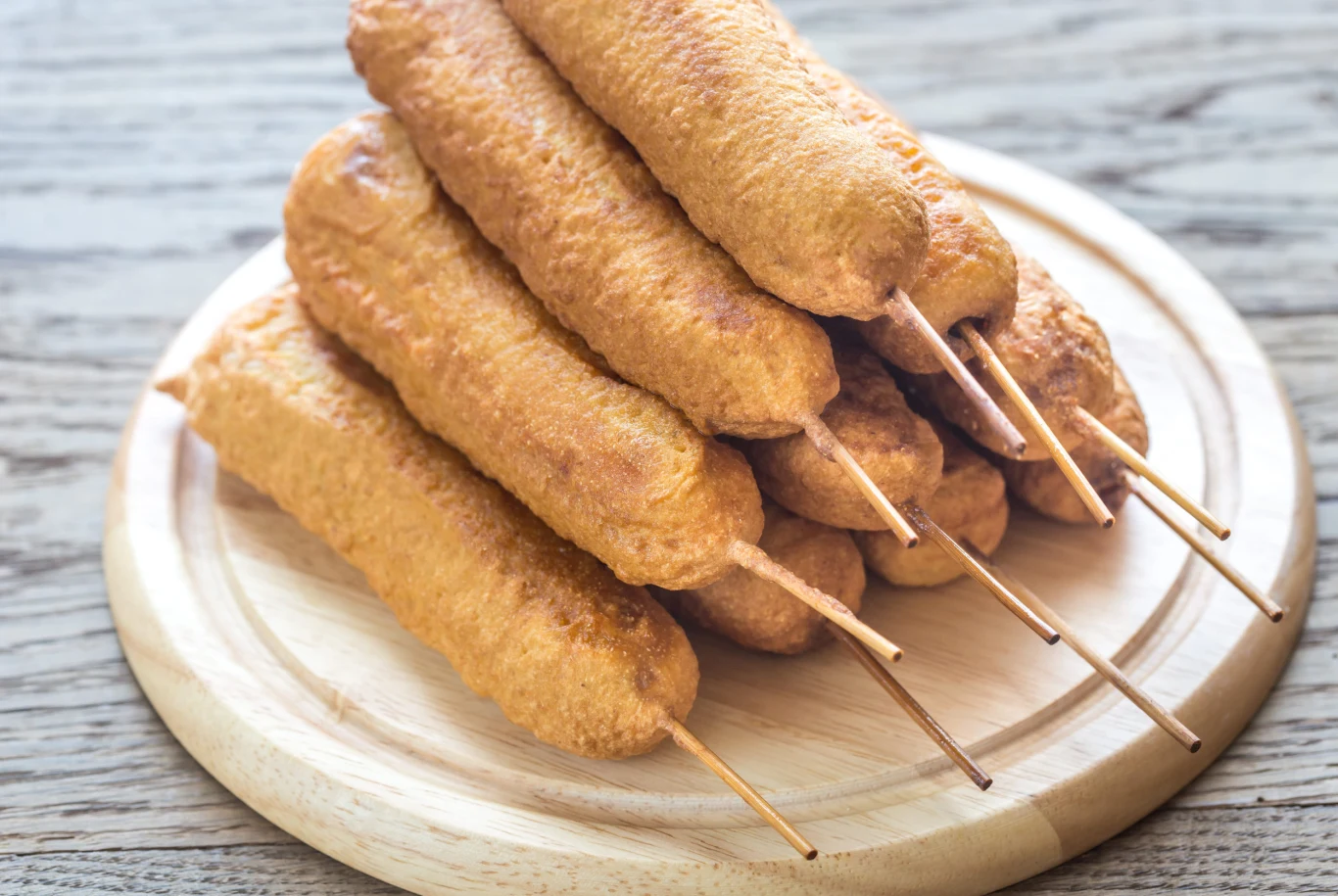Przepyszne parówki w cieście ziemniaczanym z mozzarellą. Taką przekąską nie pogardzo nawet wybredny teść Koreańskie corn dogi z parówką i mozzarellą na patykach, ułożone na drewnianej desce.