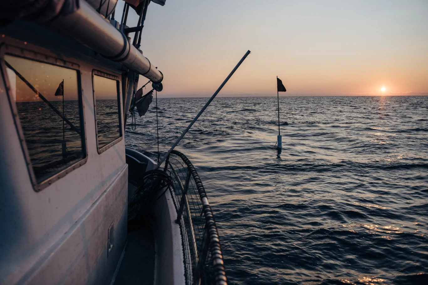 Ocean Winds wybuduje na Bałtyku 26 turbin wiatrowych. Wytworzą one energię dla ok. 500 tys. gospodarstw domowych w Polsce Burta łodzi rybackiej na tle spokojnego morza, pojedyncza boja z flagą na wodzie i słońce zachodzące przy horyzoncie.