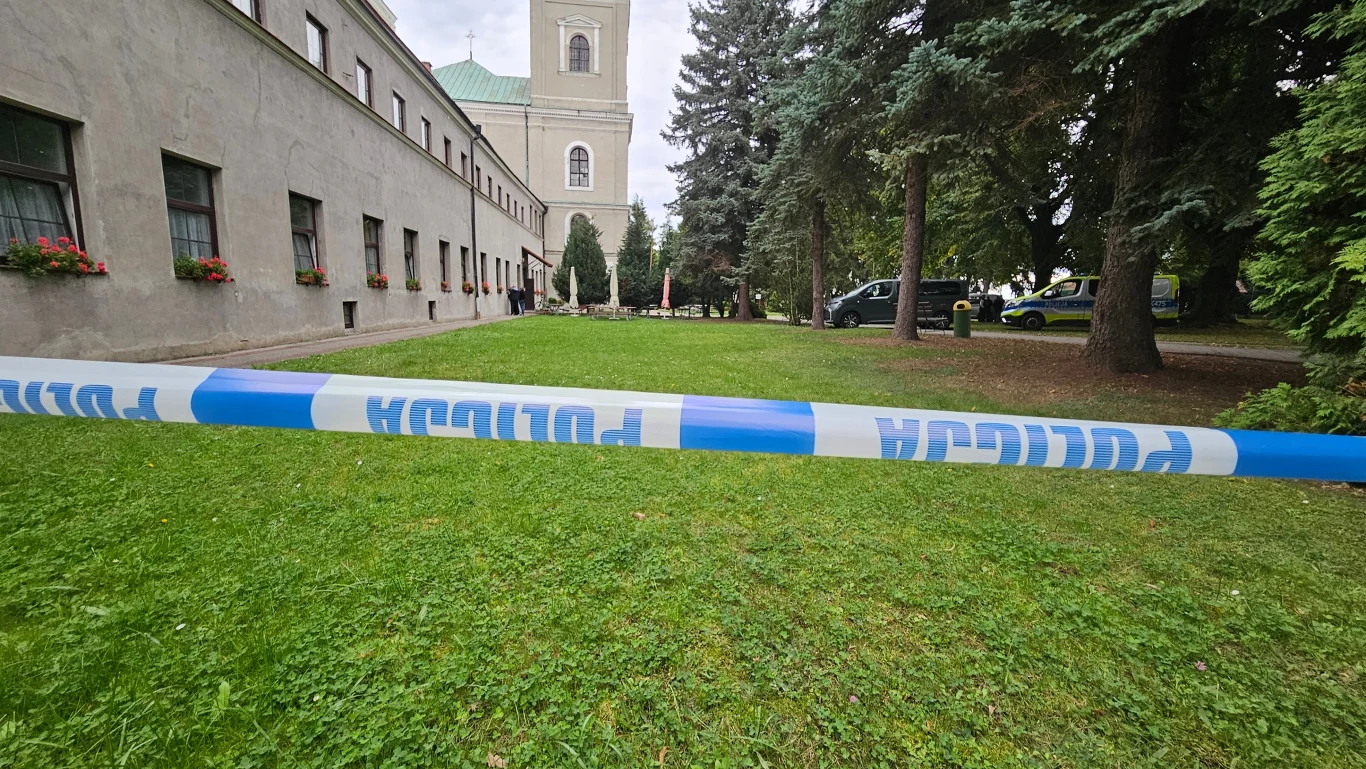 Teren ogrodzony taśmą policyjną, w tle budynek z wieżą oraz kilka zaparkowanych radiowozów w otoczeniu drzew.
