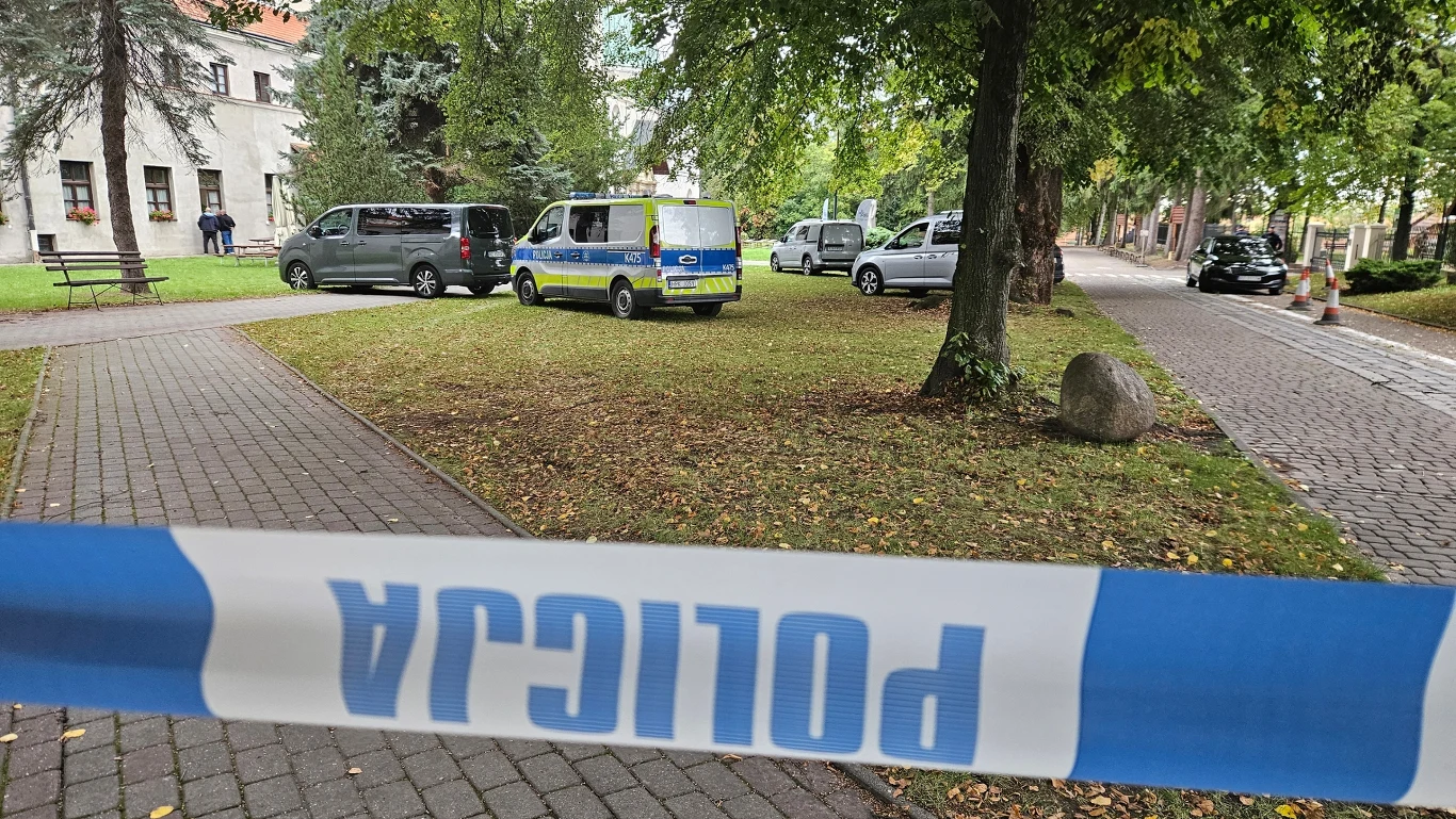 Atak nożownika w Jarosławskim Opactwie Policyjna taśma odgradzająca teren, kilka radiowozów zaparkowanych na trawie w pobliżu chodnika, funkcjonariusze pracujący w otoczeniu drzew i budynku.