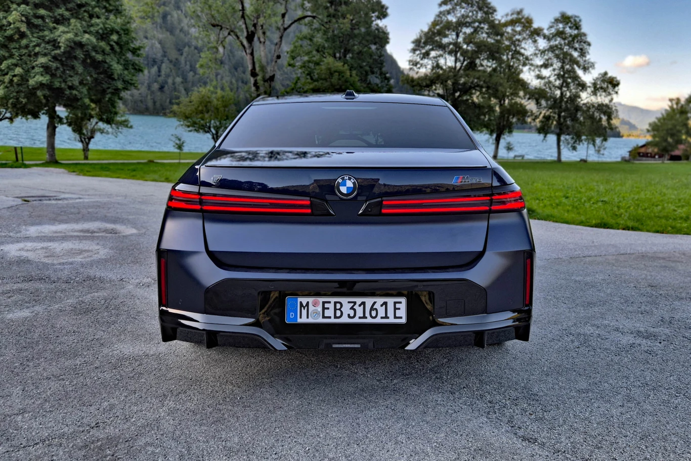 Tylne światła BMW serii 7 są bardzo szerokie, tak jak w innych modelach Neue Klasse, ale mają bardzo prosty i stonowany kształt Tylny widok luksusowego samochodu BMW z nowoczesnymi, poziomymi światłami LED i oznaczeniem modelu M760e, zaparkowanego na asfaltowej nawierzchni w otoczeniu zielonych drzew i jeziora.