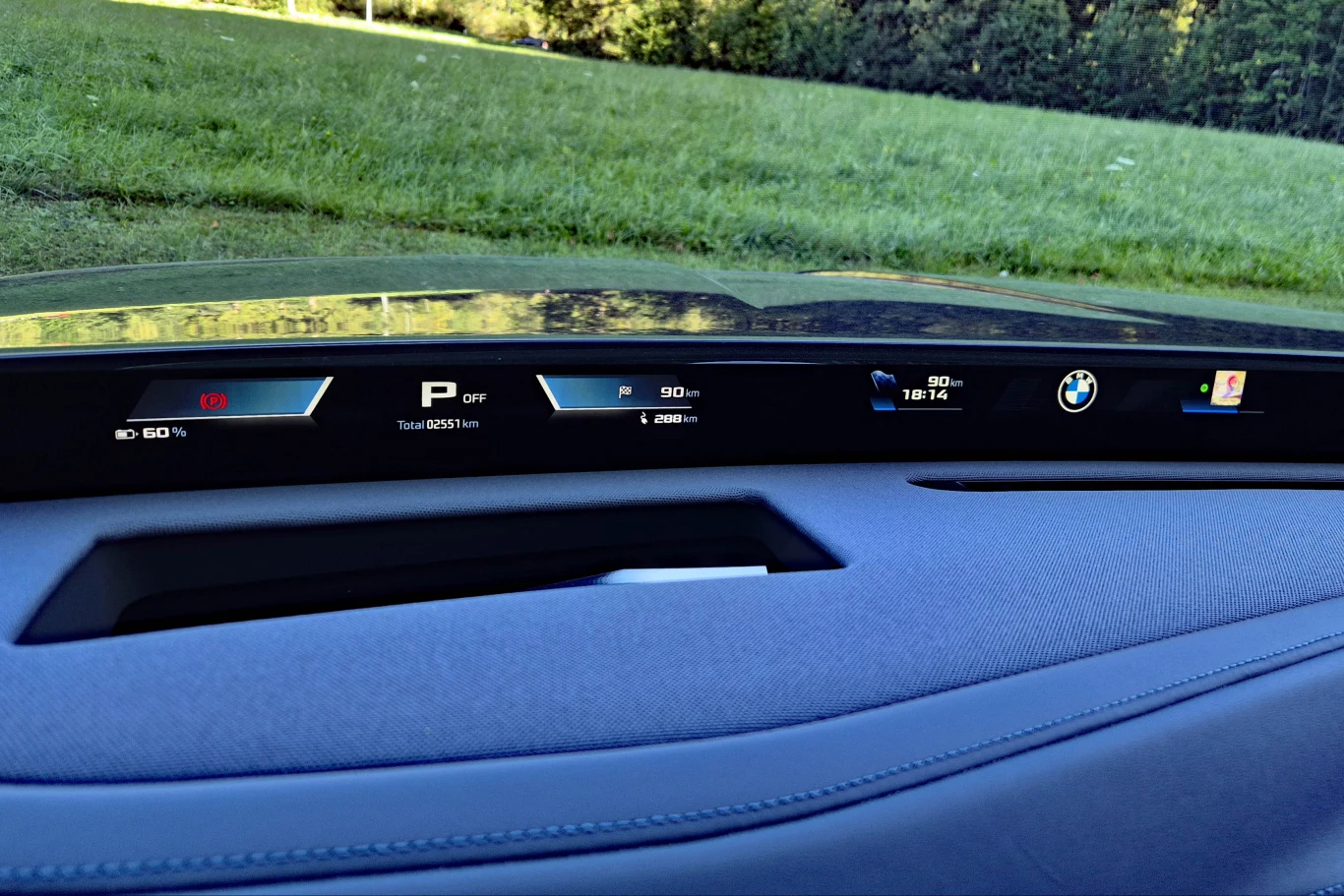 BMW Panoramic iDrive działa na zasadzie projektora, wyświetlającego wskazania na wyczernionym podszybiu Cyfrowy panel kokpitu nowoczesnego samochodu, wyświetlający informacje o prędkości, trybie jazdy, poziomie naładowania akumulatora oraz logo marki BMW.