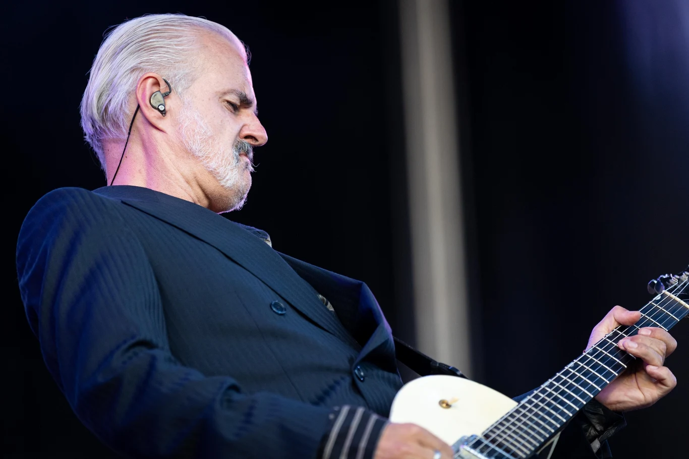 Mężczyzna z siwymi włosami i brodą grający na białej gitarze elektrycznej, ubrany w ciemny garnitur, skupiony podczas występu na scenie.