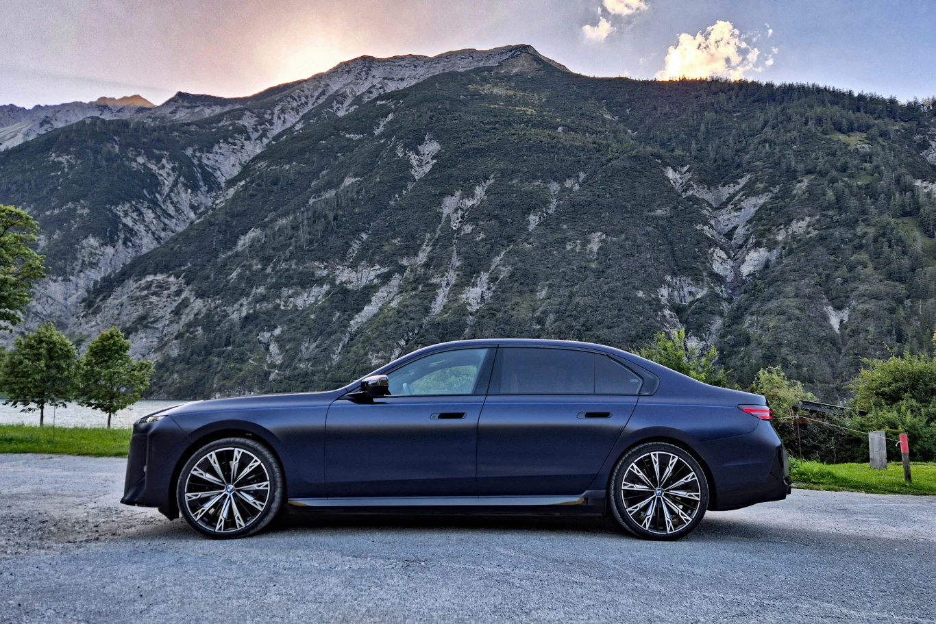 BMW serii 7 niezmiennie dostępne jest tylko w długiej wersji, mierzącej 5395 mm Luksusowy, ciemnoniebieski sedan sportowy stojący na tle wysokich, zielonych gór porośniętych lasem, z widocznymi charakterystycznymi felgami i elegancką sylwetką samochodu.