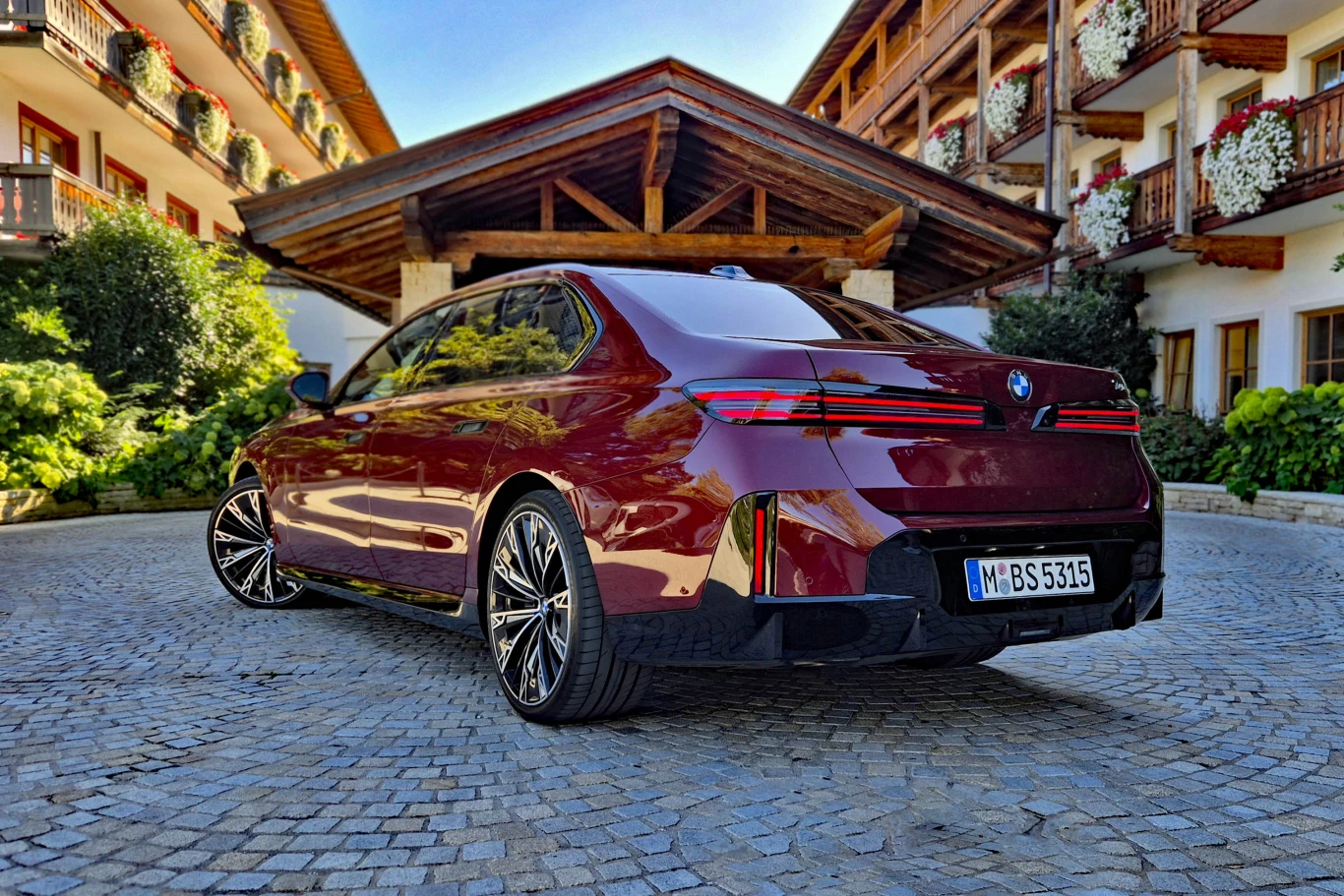 Również z tyłu BMW 740 nie zdradza, że mamy do czynienia z wersją spalinową - rury wydechowe ukryto pod zderzakiem Nowoczesny sedan BMW w bordowym kolorze zaparkowany na brukowanym dziedzińcu, otoczony drewnianą architekturą z balkonami i kwiatami.