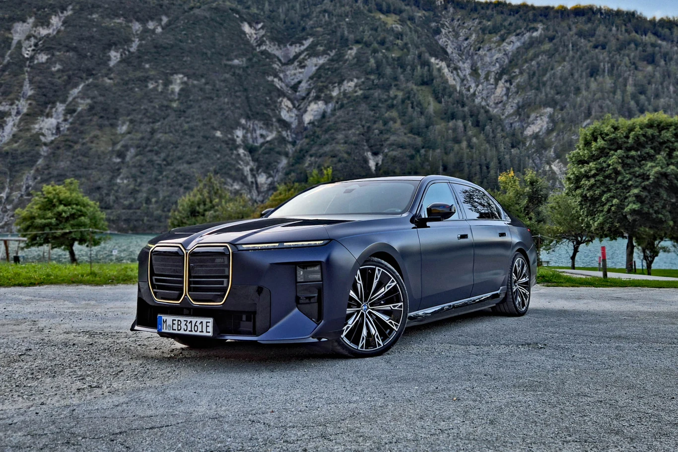 BMW serii 7 Granatowy luksusowy sedan BMW z charakterystycznym dużym grillem, sportowymi felgami i futurystycznym designem, ustawiony na tle górskiego krajobrazu z jeziorem i zielonymi drzewami.