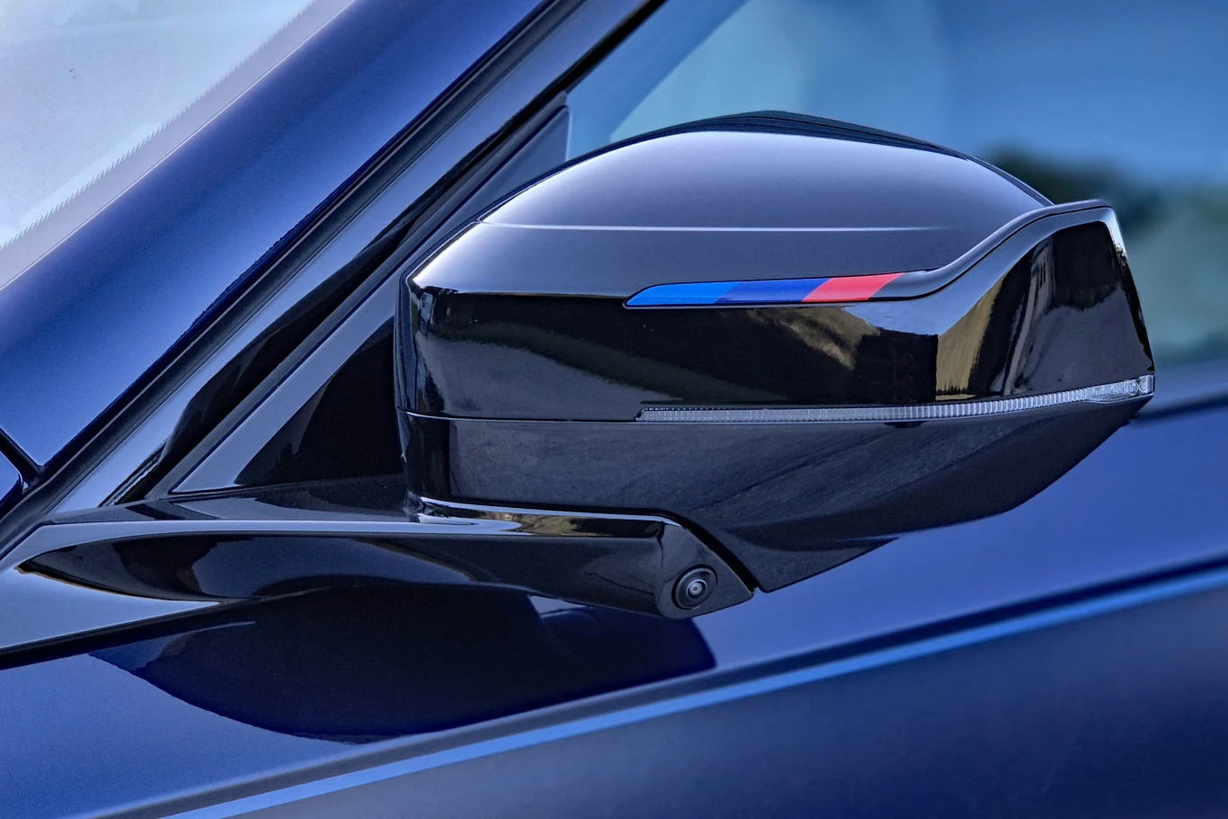 BMW i7 M70 Czarny lusterko boczne samochodu z charakterystycznym paskiem w niebiesko-czerwonych barwach, osadzone na lakierowanej karoserii w odcieniu granatu.