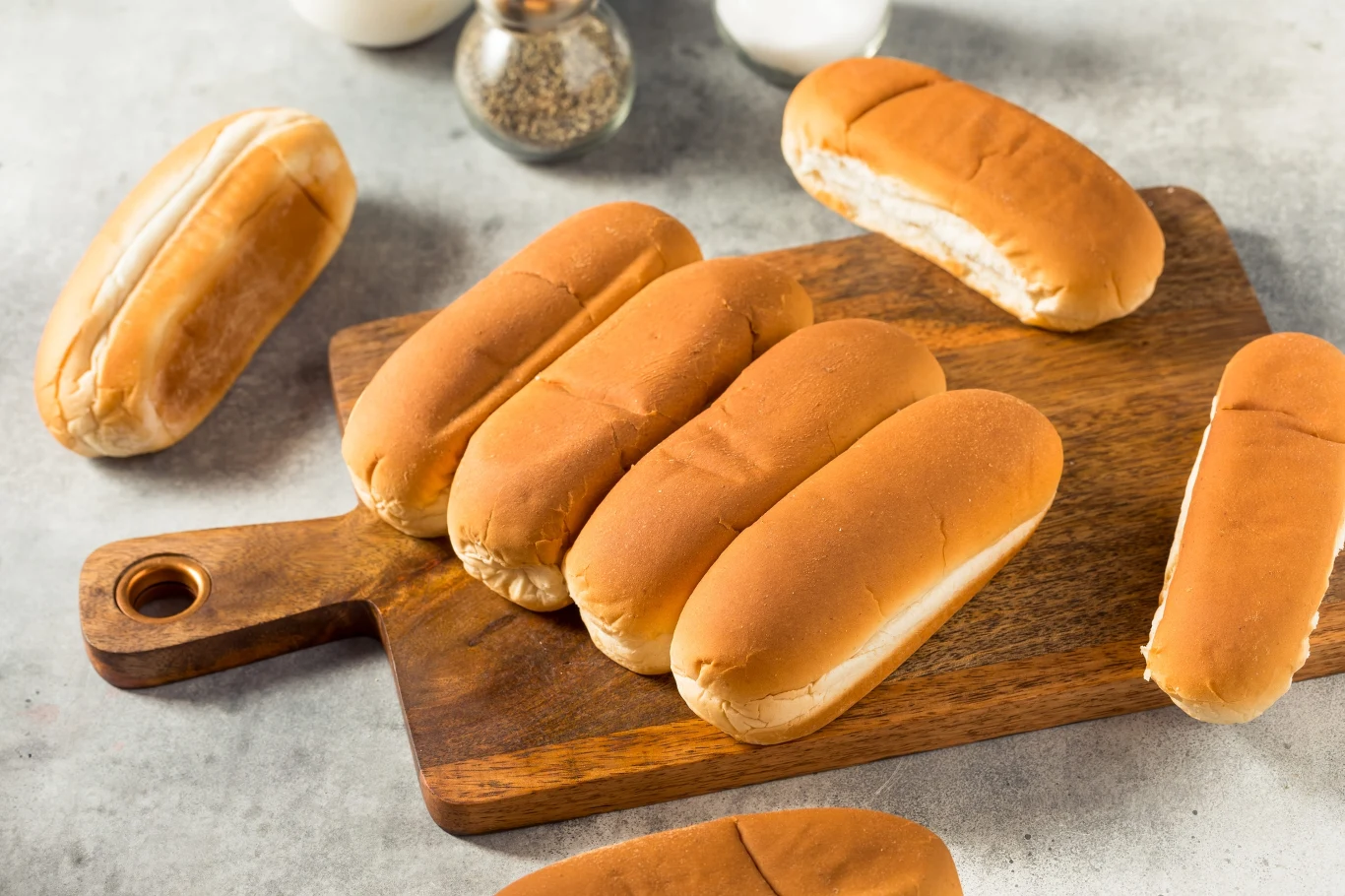 Kont Skontrolowano 80 podmiotów, które wprowadzały do obrotu asortyment opakowanych wyrobów piekarskich Pięć świeżych, podłużnych bułek do hot dogów ułożonych na drewnianej desce do krojenia, wokół niej rozmieszczone są pojedyncze bułki.