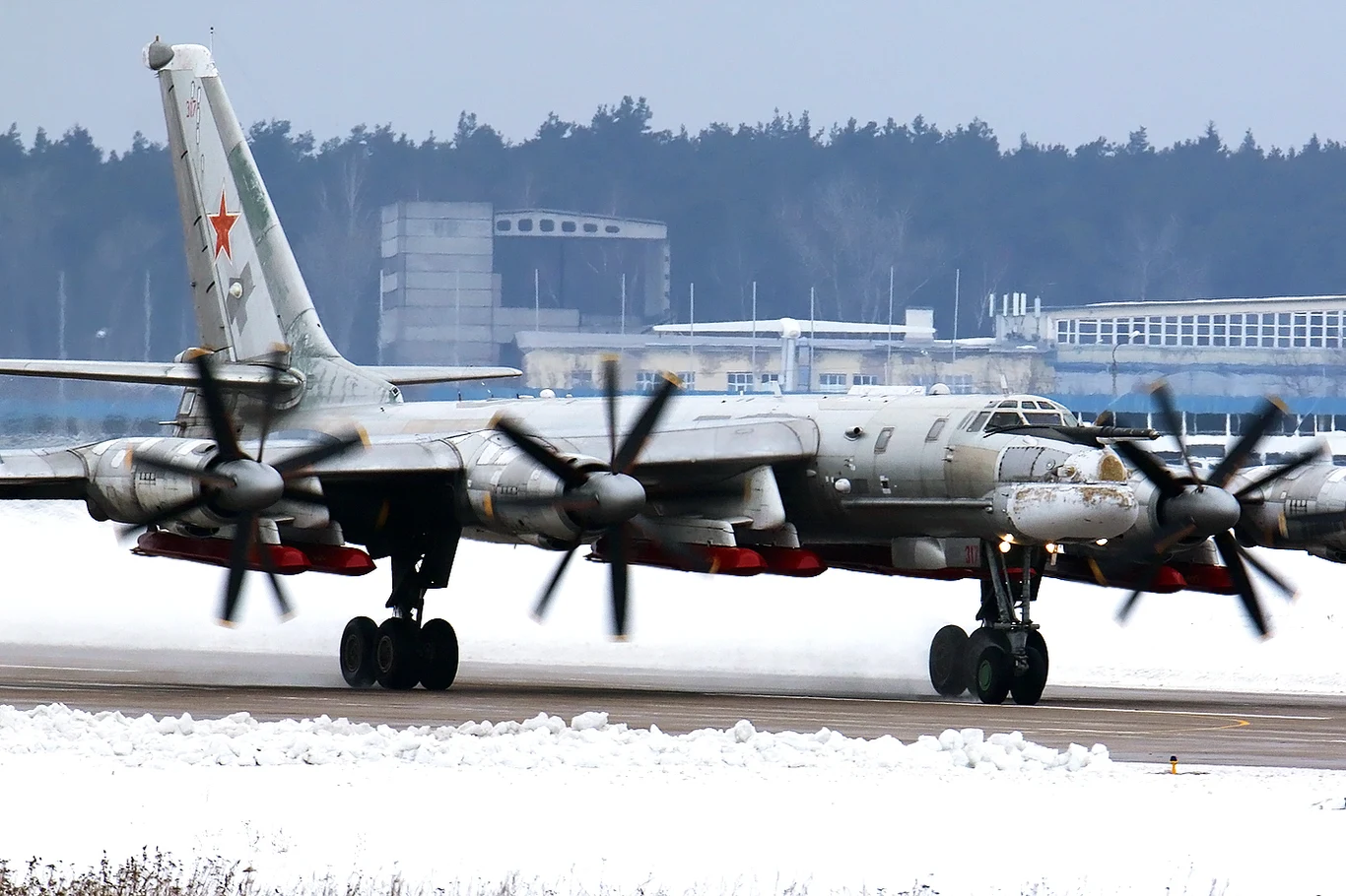 Rosyjski bombowiec strategiczny Tu-95MS z podwieszonymi pociskami Raduga Ch-101/102, które mogą przenosić ładunek atomowy. Samolot wojskowy z napędem turbośmigłowym, w barwach sił rosyjskich, kołuje po zaśnieżonym pasie startowym, w tle zabudowania lotniskowe i las.