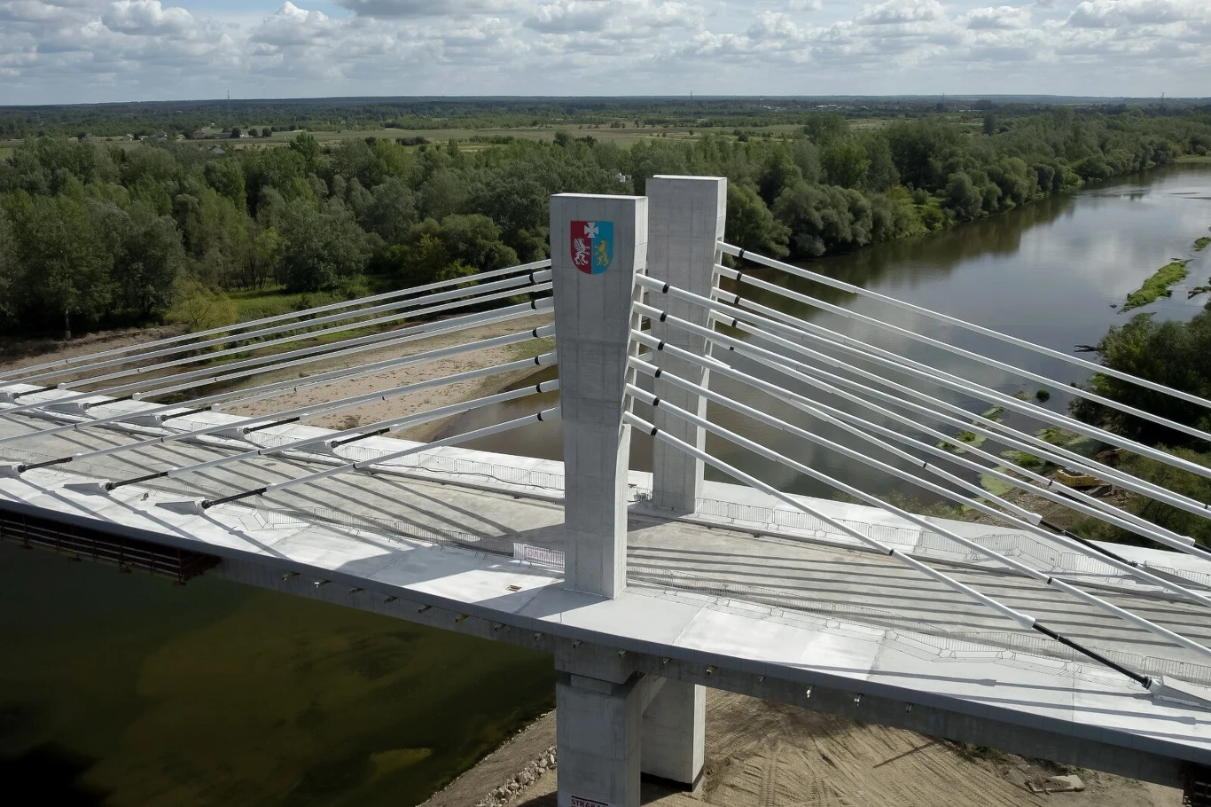 Nowoczesny most podwieszany z betonowymi pylony i stalowymi linami rozciągniętymi nad szeroką rzeką, otoczony zielonym lasem po obu stronach brzegu.
