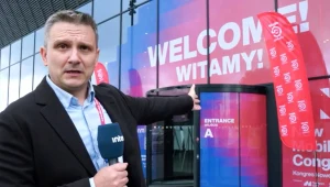 Mężczyzna w garniturze trzymający mikrofon z logotypem stacji telewizyjnej wskazuje ręką na wejście do budynku oznaczone dużym napisem powitalnym oraz flagami wydarzenia.