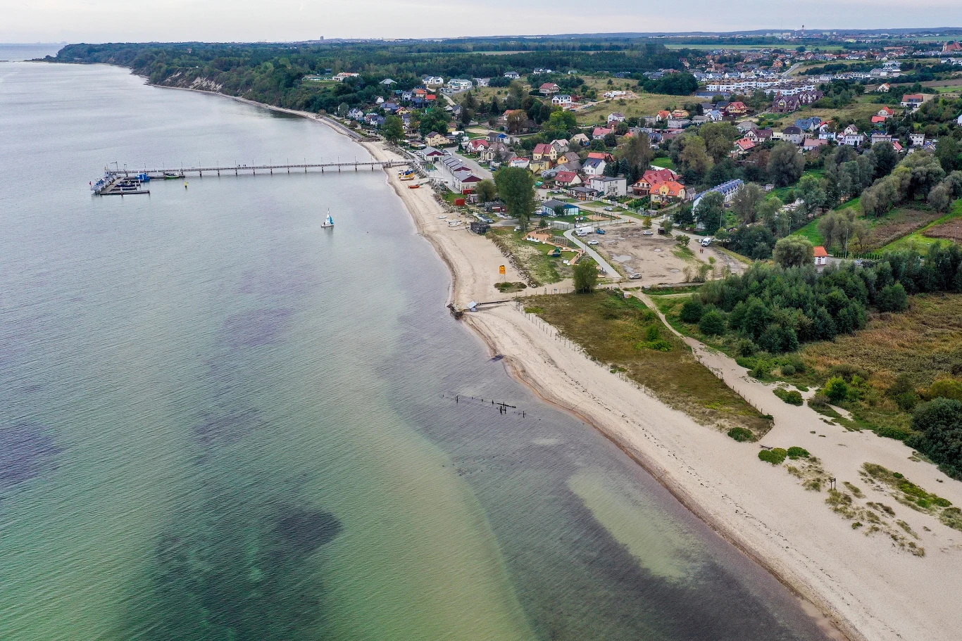 Piaszczysta plaża z łagodnym zejściem do morza, długie molo wychodzące w głąb wody oraz otoczona zielenią zabudowa nadmorskiego miasteczka.