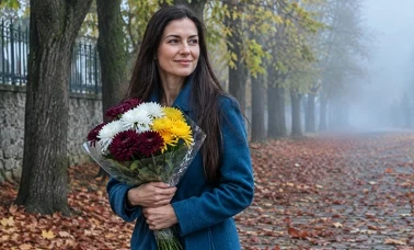 Kobieta z bukietem sztucznych chryzantem idzie mglistą jesienną aleją wśród opadłych liści 