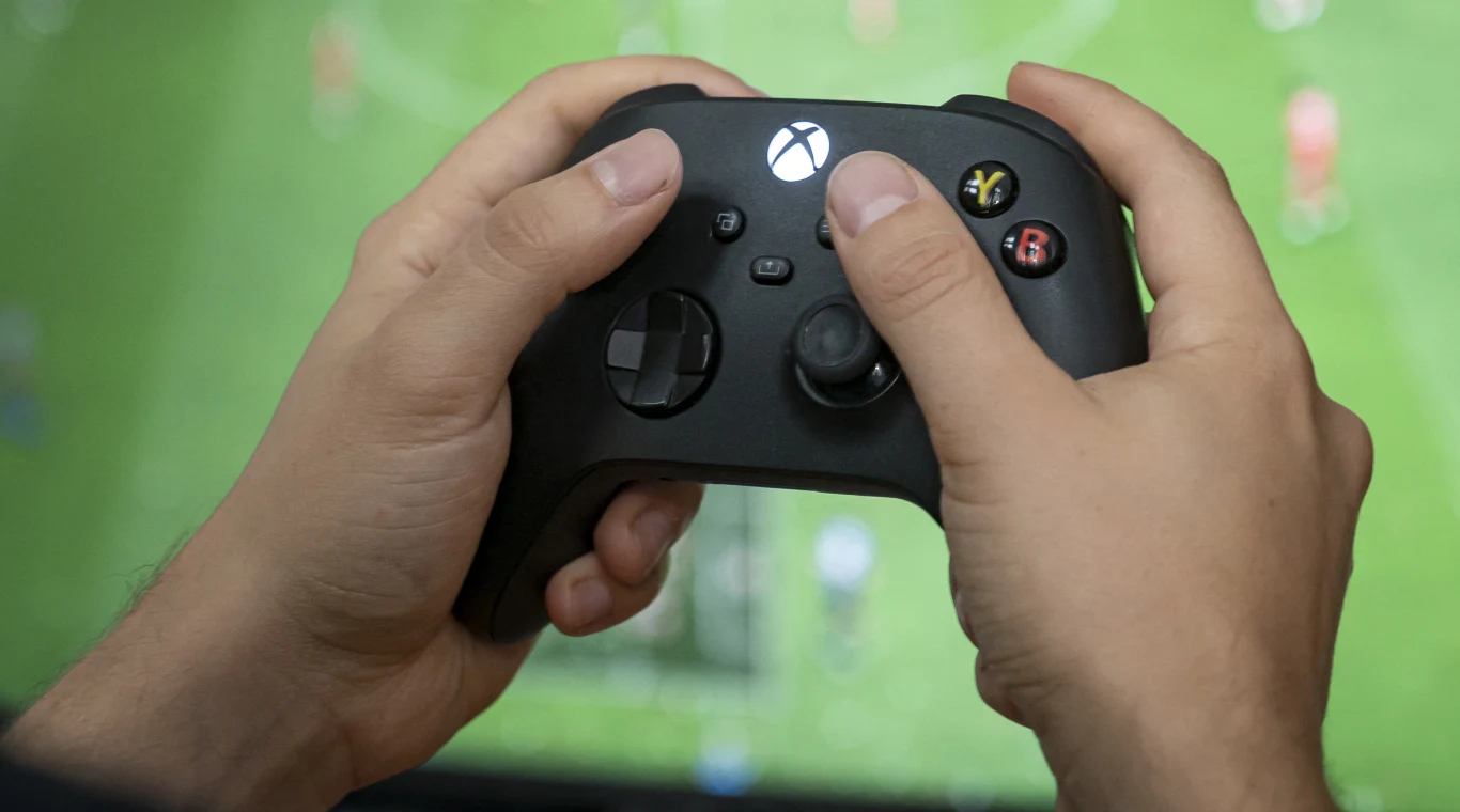 Patent Microsoftu analizuje obraz i dźwięk z gry pod reklamy Ręce z czarnym kontrolerem Xbox, w tle ekran z grą piłkarską, ilustracja mechanizmu patentu Microsoftu.