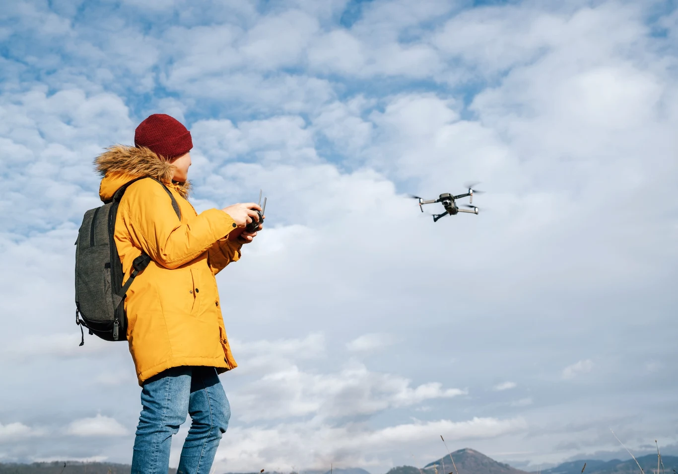 Gmina Niepołomice uzupełnia sprzęt w szkole. Zamawia m.in. drony. Osoba w żółtej kurtce i bordowej czapce steruje dronem unoszącym się na tle nieba z chmurami.