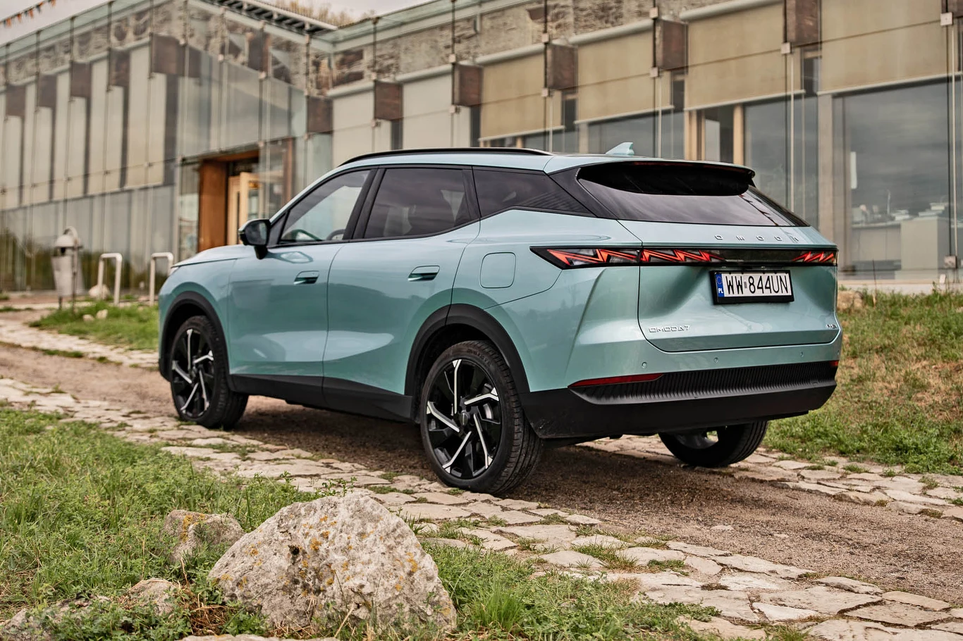 Lakier Mist Green sprawia, że Omoda 7 tym bardziej wyróżnia się na ulicy Nowoczesny, jasnoniebieski SUV z czarnym dachem i ciemnymi felgami stoi na nieutwardzonej drodze przy budynku o szklanej elewacji.