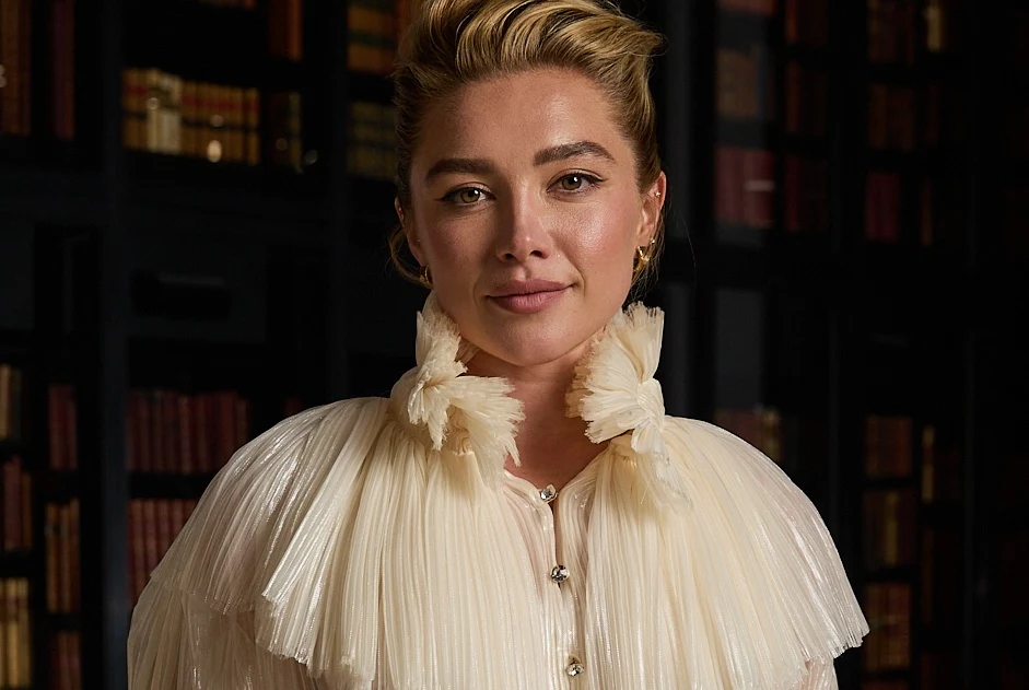 Florence Pugh Florence Pugh w kremowej, plisowanej bluzce ze spinkami, stoi na tle regału z książkami.