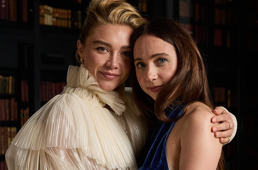 Florence Pugh i Zoe Kazan Dwie uśmiechnięte kobiety obejmują się, jedna ubrana w jasną, plisowaną suknię z falbaną przy szyi, druga w ciemnoniebieską kreację na ramiączkach, stoją na tle półek z książkami.