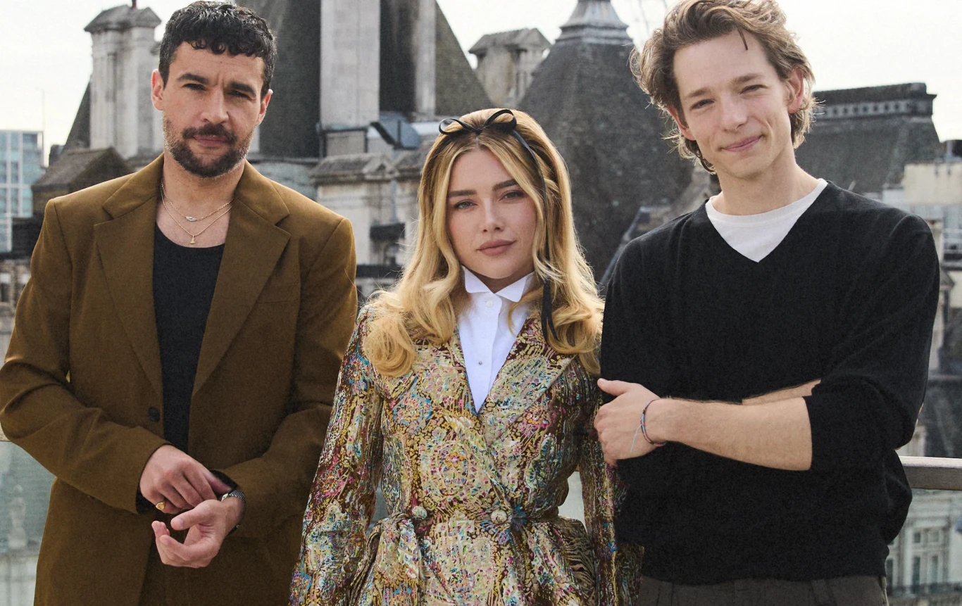 Christopher Abbott, Florence Pugh, Mike Faist Trzy osoby stojące razem na tle miejskich dachów; mężczyzna po lewej w brązowej marynarce, kobieta pośrodku w kolorowej sukience i mężczyzna po prawej w czarnym swetrze z podwiniętymi rękawami.