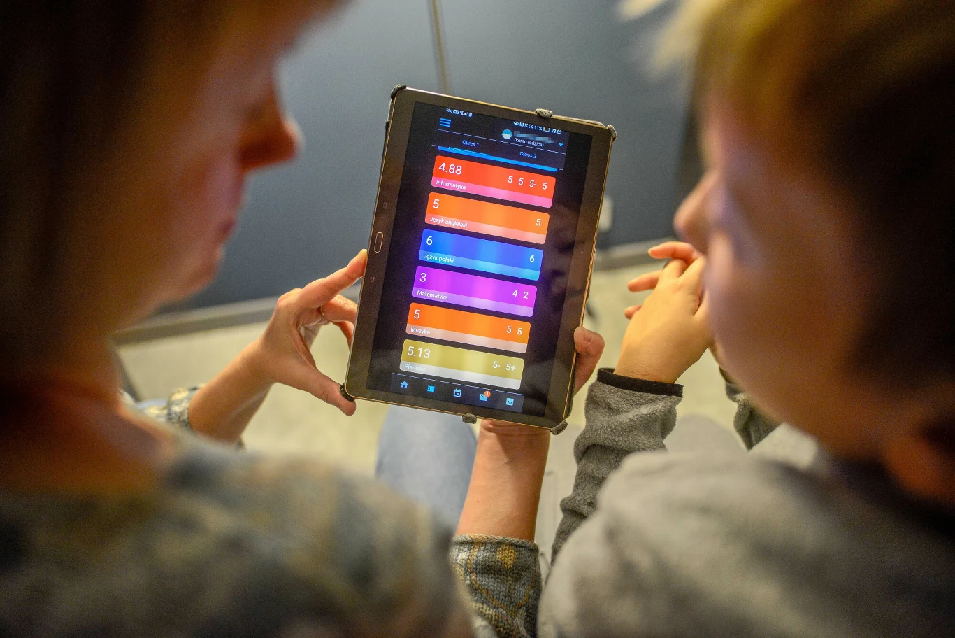 Tablet z kolorową aplikacją edukacyjną wyświetlający działania matematyczne trzymany przez dorosłą osobę, obok siedzi dziecko uważnie patrzące na ekran.