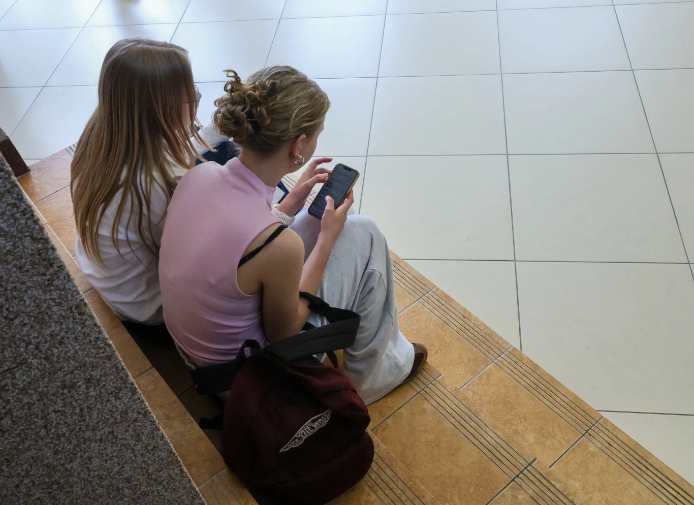 Dwie dziewczyny siedzące na schodach, skupione na telefonach komórkowych, z plecakiem leżącym obok.