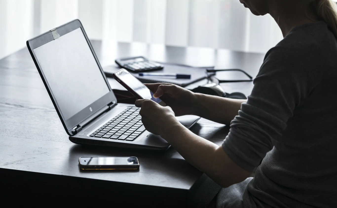 Osoba siedząca przy biurku korzysta z laptopa i trzyma w ręku smartfon, obok na blacie leży drugi telefon oraz kalkulator.