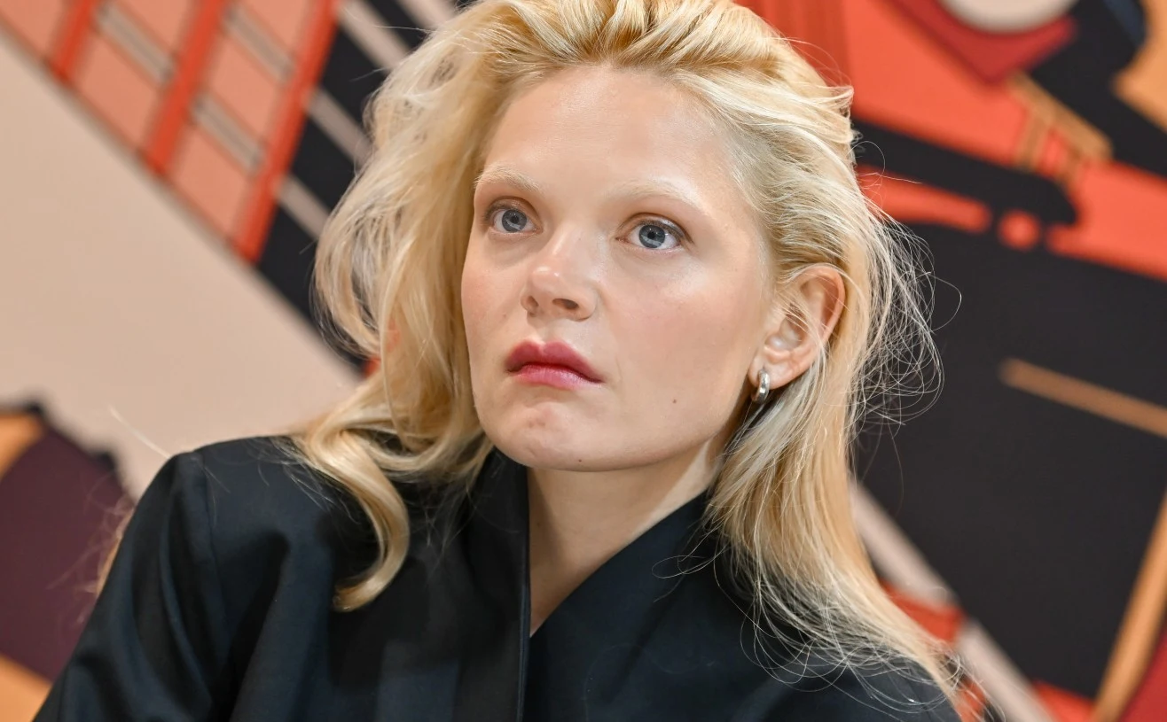 Sandra Drzymalska Kobieta o blond włosach i jasnej cerze w ciemnej marynarce, siedząca na tle abstrakcyjnej grafiki w ciepłych kolorach.