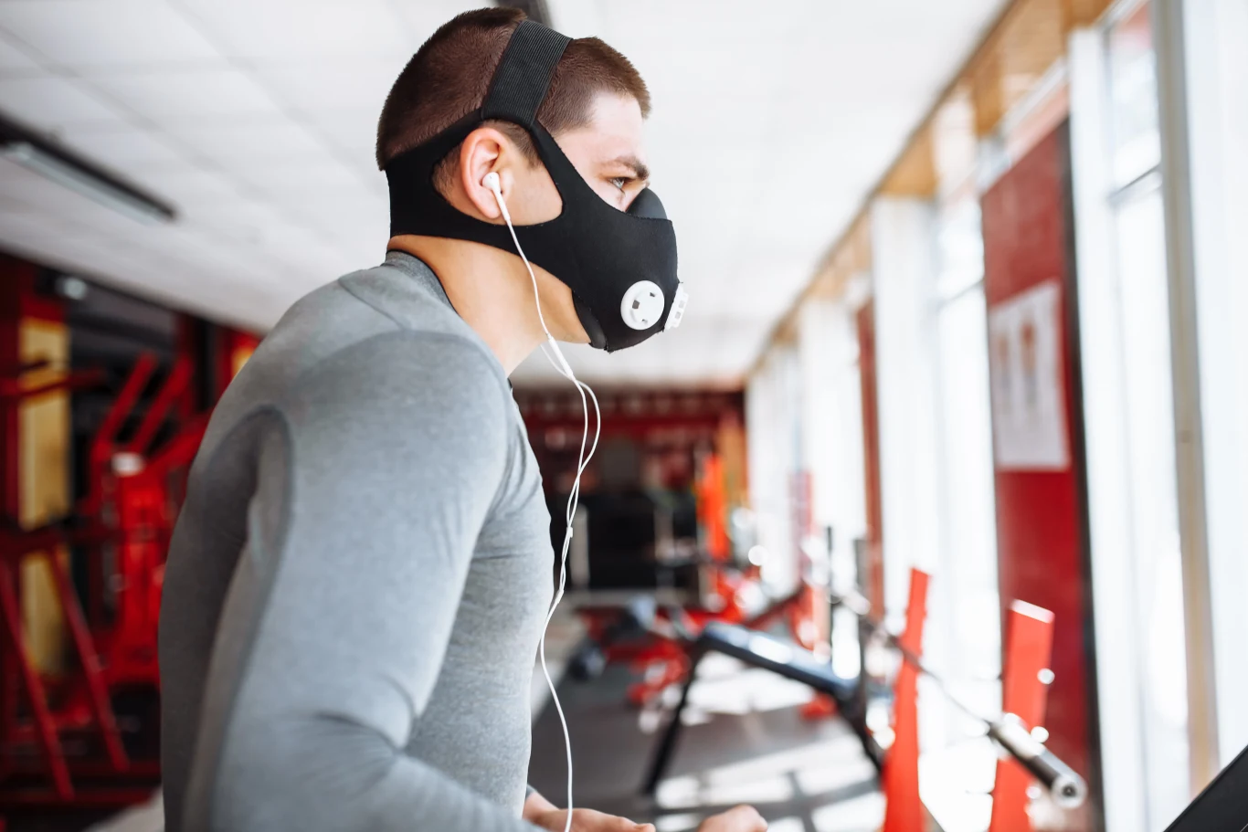 Mężczyzna w sportowej odzieży biegnący na bieżni, na twarzy maska treningowa i słuchawki, wnętrze siłowni z dużą ilością światła dziennego.