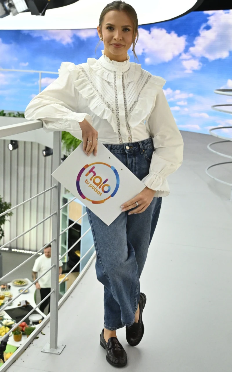 Kobieta ubrana w białą koszulę z falbanami oraz niebieskie jeansy, stojąca na schodach z planszą programu 'halo dzień dobry' na tle nowoczesnego studia telewizyjnego.