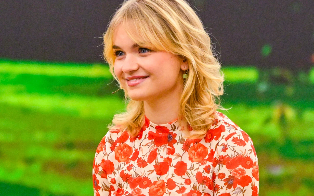 Marcelina "Marcycha" Ruczyńska Młoda kobieta o blond włosach w czerwonej sukience w kwiaty, uśmiechnięta, na tle rozmytego zielonego pejzażu.