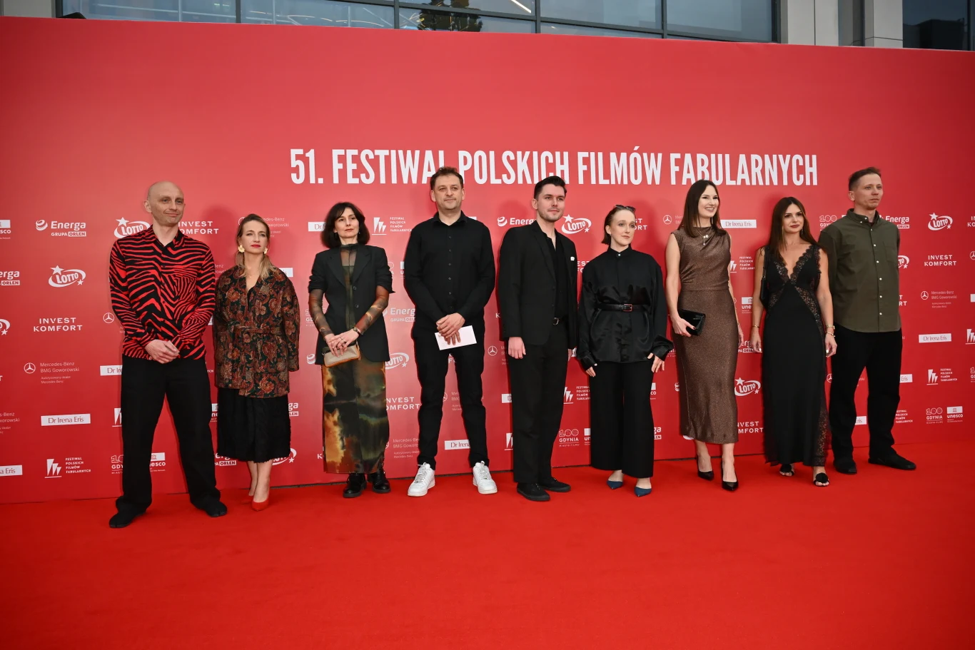 Grupa dziewięciu osób ubranych w eleganckie stroje pozuje na czerwonym dywanie na tle ściany z logo 51. Festiwalu Polskich Filmów Fabularnych.