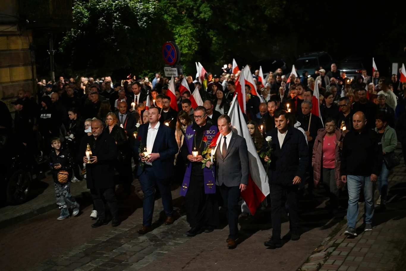 Marsz Ciszy w Jarosławiu Tłum ludzi idący nocą ulicą z flagami Polski, świecami oraz zniczami; na przedzie grupa mężczyzn w garniturach i duchowny w fioletowej stule.