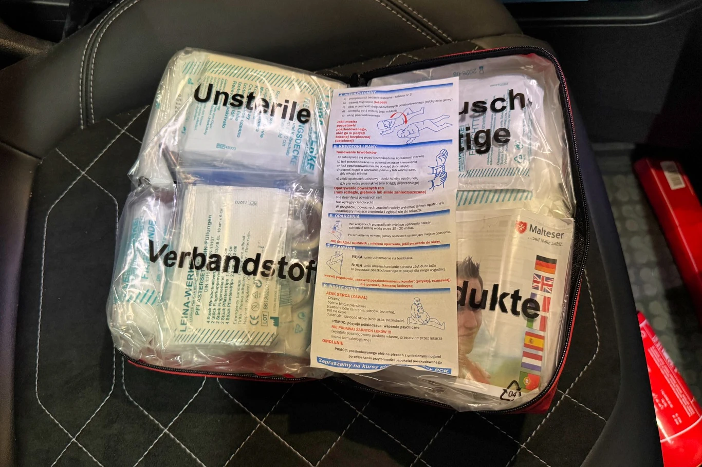 W apteczce samochodowej powinny znaleźć się przede wszystkim materiały opatrunkowe, rękawiczki, nożyczki oraz koc ratunkowy. Leków lepiej w niej nie przechowywać. Zestaw pierwszej pomocy z widocznymi opatrunkami i instrukcją udzielania pomocy, umieszczony na czarnym fotelu samochodowym.
