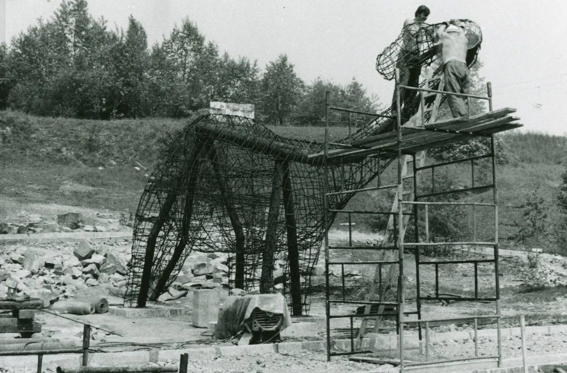 Budowa "Kotliny Dinozaurów". Lata 70. Szkielet dużej konstrukcji przypominającej smoka budowany przez dwóch pracowników na rusztowaniu, otoczony materiałami budowlanymi i narzędziami na tle porośniętego drzewami pagórka.