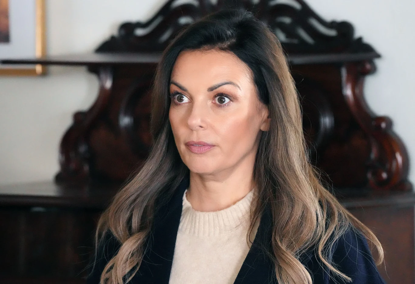 Kasia zgodzi się pośredniczyć w sprzedawaniu warzyw i owoców Janki Kobieta o długich, brązowych włosach i wyrazistych brwiach, ubrana w jasny sweter i ciemną marynarkę, stoi na tle ozdobnego, drewnianego mebla.