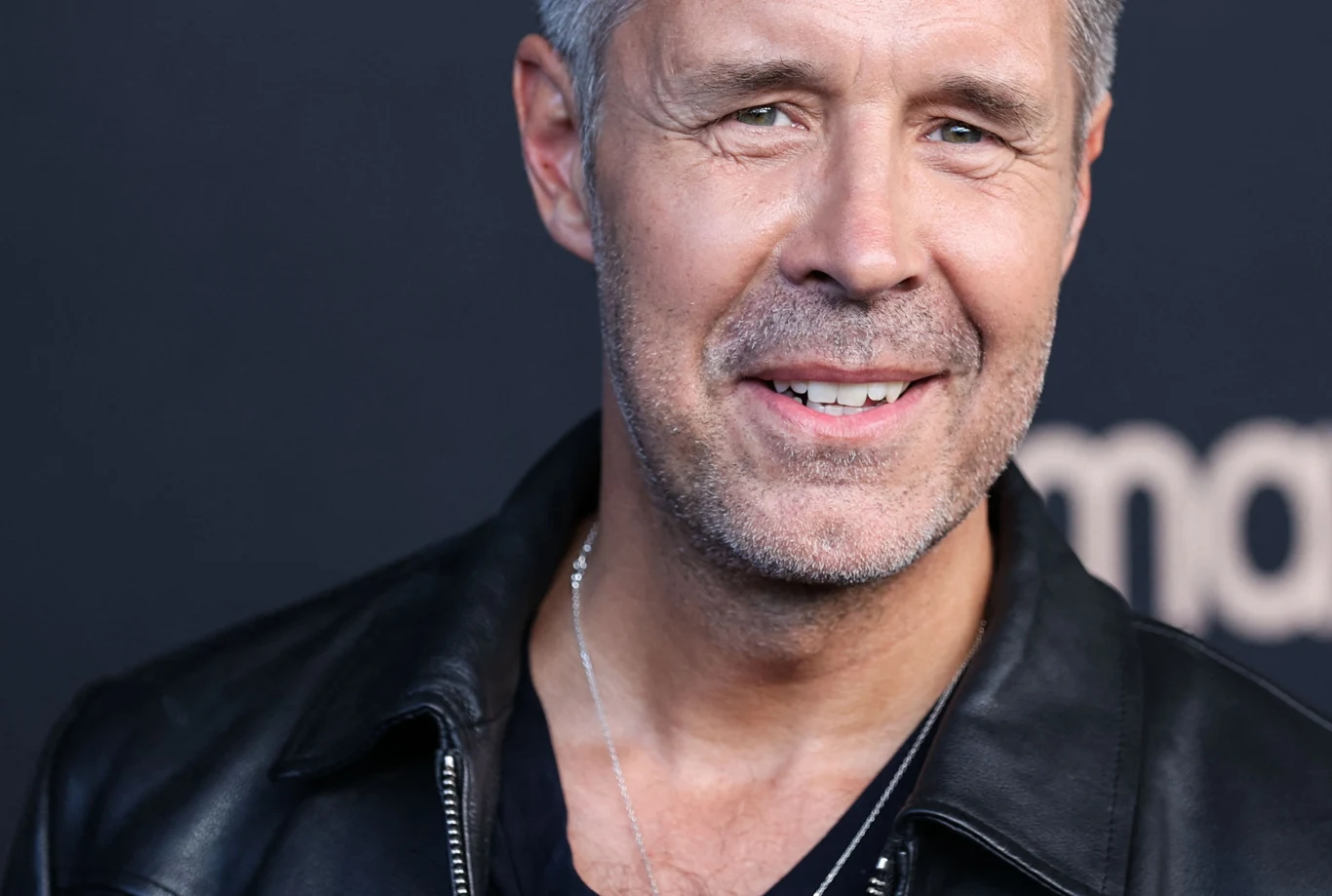 Paddy Considine Paddy Considine w czarnej skórzanej kurtce i t-shircie, srebrny łańcuszek, ciemne tło z napisem.