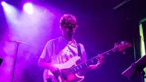 Młody gitarzysta grający na gitarze basowej podczas koncertu, otoczony fioletowym światłem scenicznym.