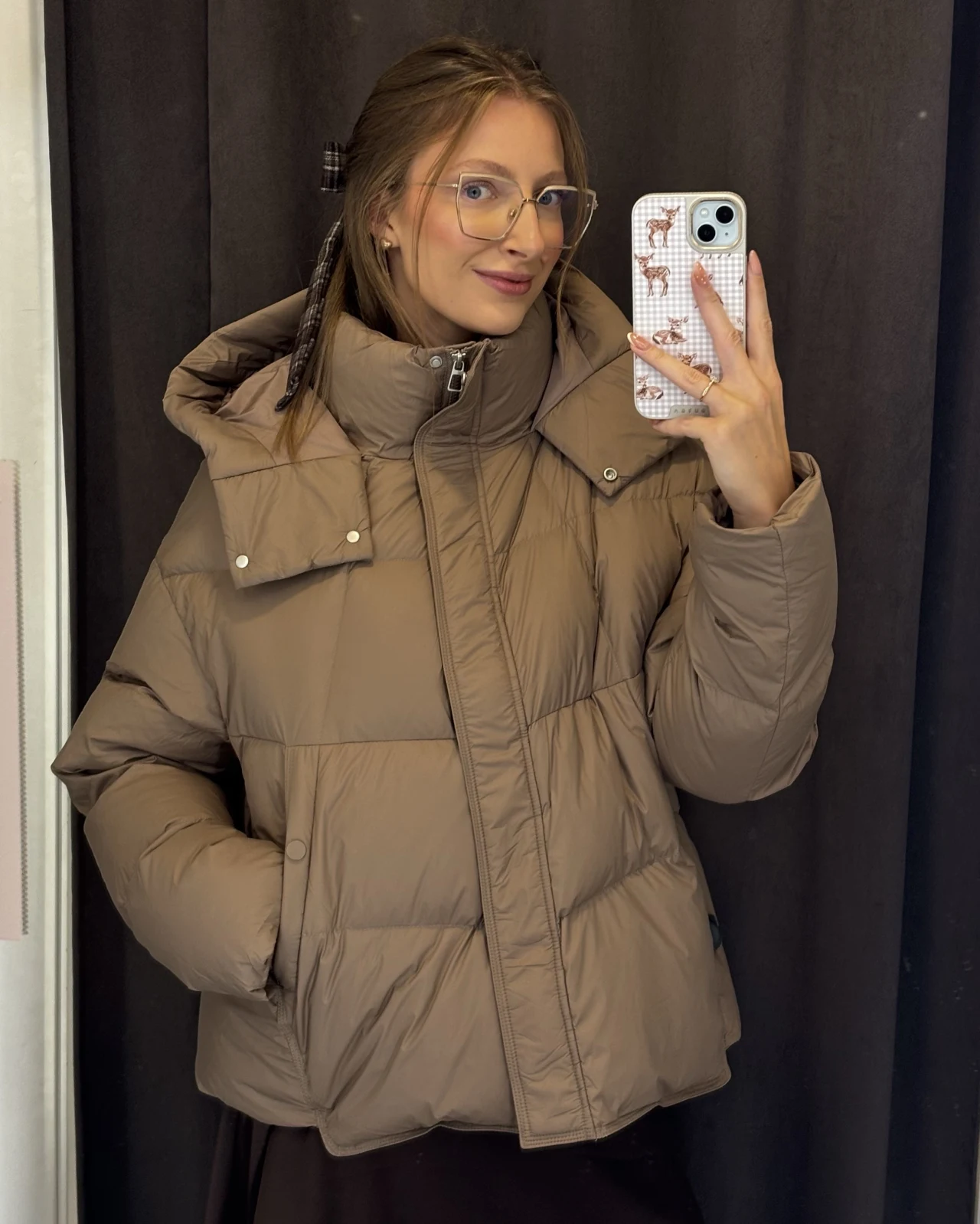 Kurtka z naturalnym puchem z Mohito. Cena: 399 zł Uśmiechnięta młoda kobieta w okularach i beżowej, pikowanej kurtce puchowej z podniesionym kołnierzem robi selfie w lustrze smartfonem w etui z nadrukiem zwierzęcym.