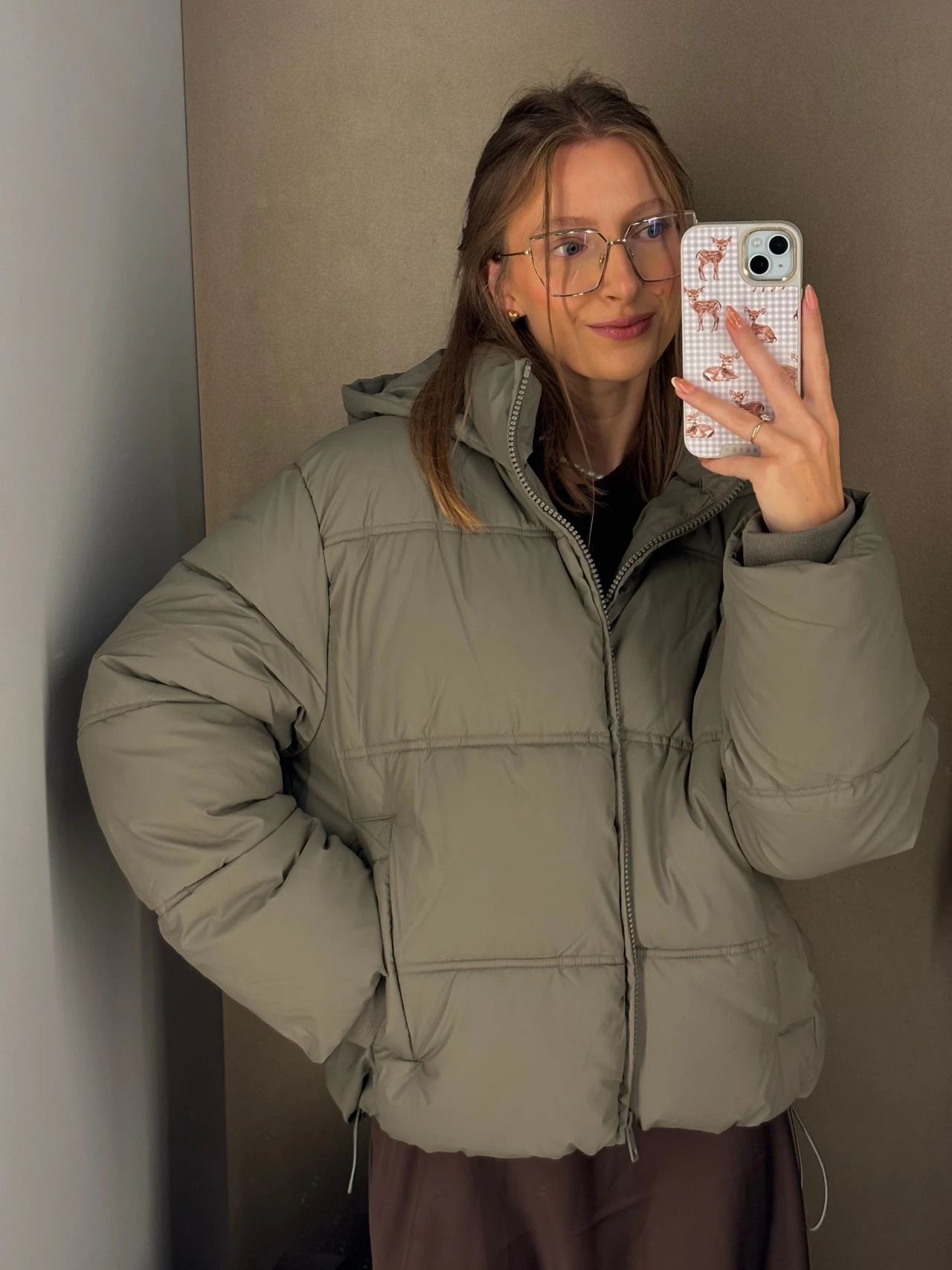 Przegląd puchowych kurtek z sieciówek Kobieta w okularach i jasnoszarej, pikowanej kurtce z kapturem trzyma smartfon i robi selfie w lustrze.