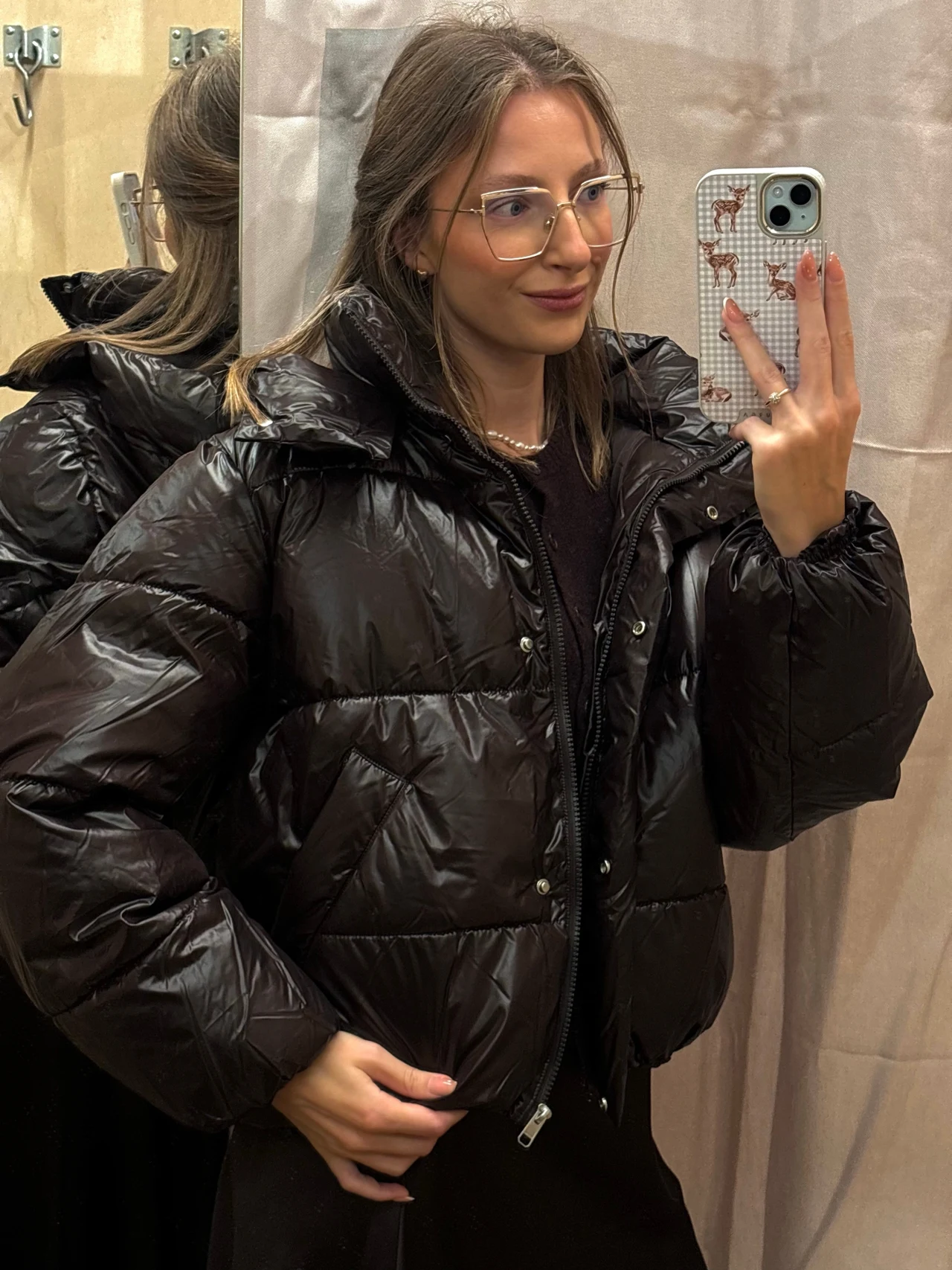 Pikowna kurtka oversize z Bershki. Cena 299 zł Młoda kobieta z długimi włosami i okularami pozuje przed lustrem, ubrana w błyszczącą, czarną, pikowaną kurtkę puchową i trzyma smartfon w ręku.