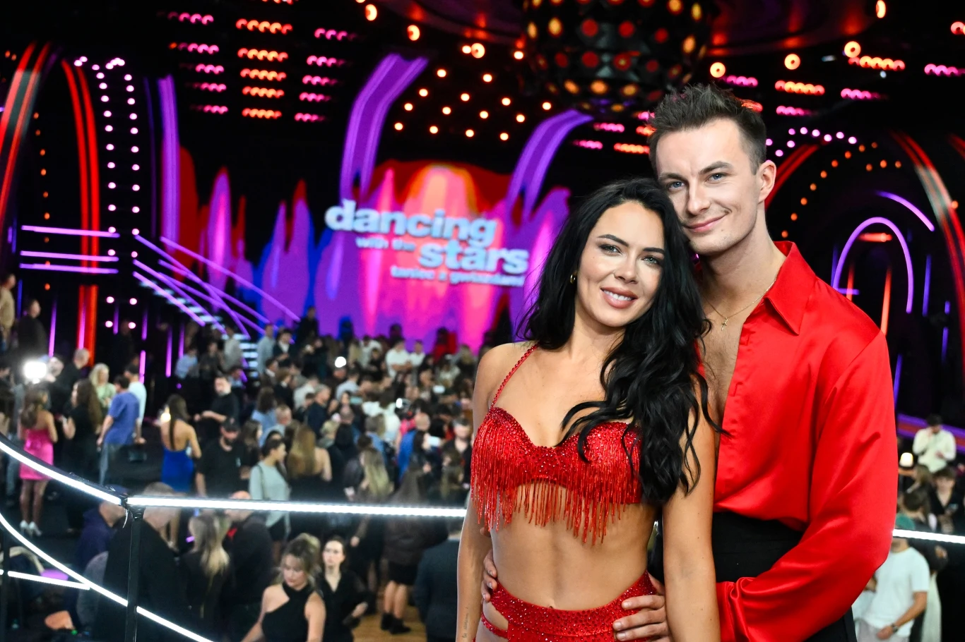 Kacper Rutka, Kaeyra Para w czerwonych, tanecznych strojach pozują razem na tle kolorowej, oświetlonej sceny programu "Dancing with the Stars", wśród tłumu widzów na parkiecie.