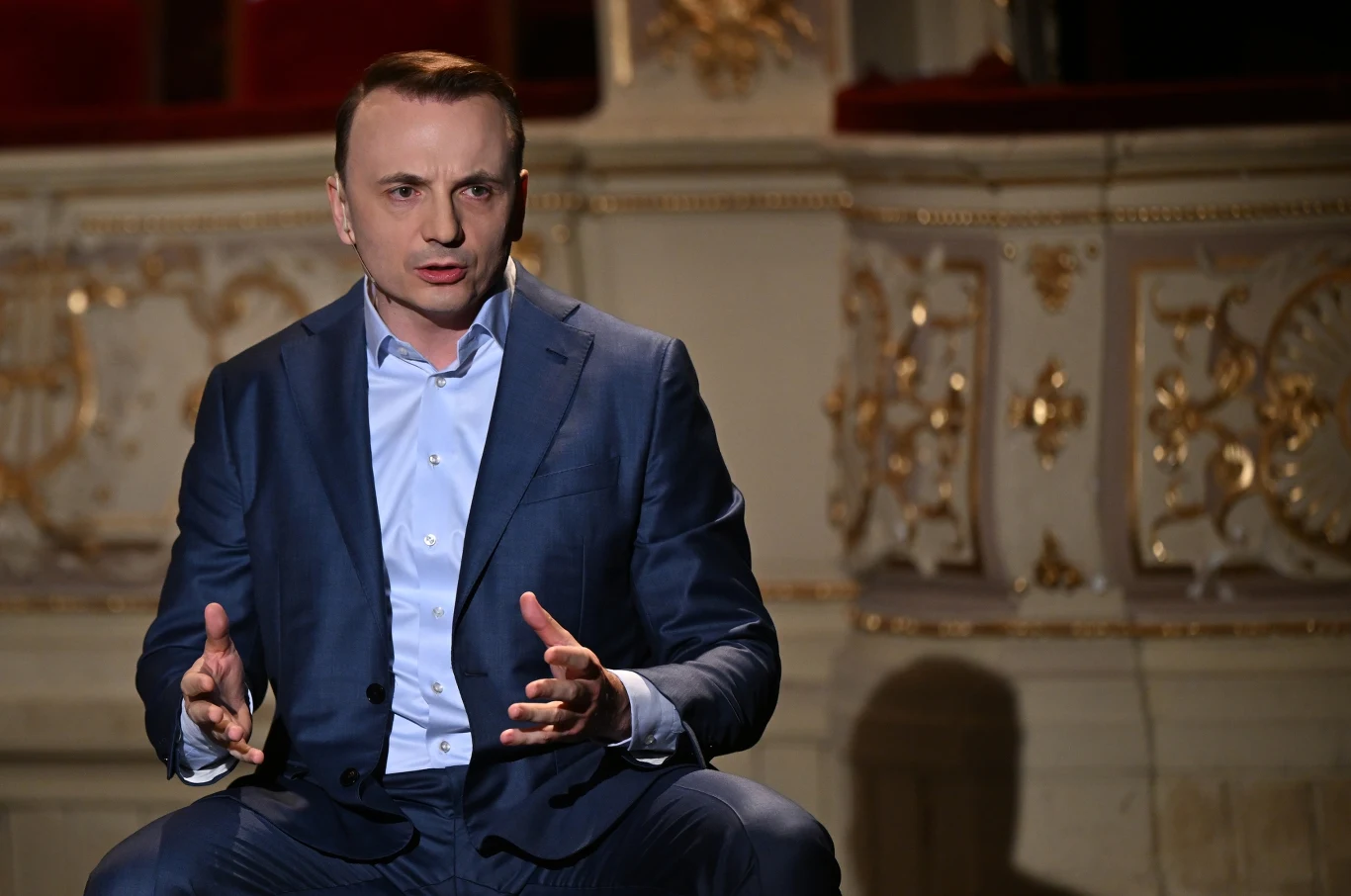 Łukasz Gibała zapowiada zmiany w krakowskim magistracie Łukasz Gibała w granatowym garniturze gestykuluje podczas wypowiedzi we wnętrzu ze złotymi detalami.