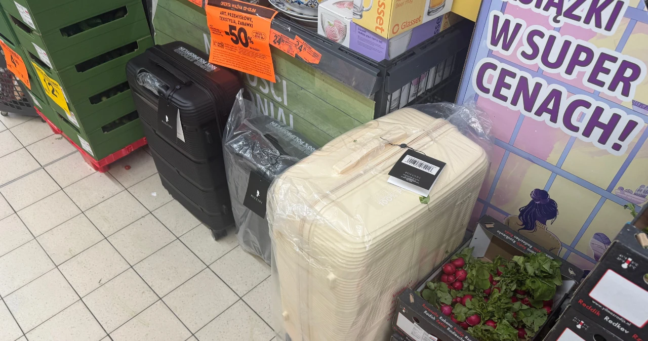 Próchnik wchodzi do Biedronki. Walizki od 139 zł to dopiero początek Dwie walizki w różnych kolorach przy regałach Biedronki obok warzyw i tabliczki promocyjnej.